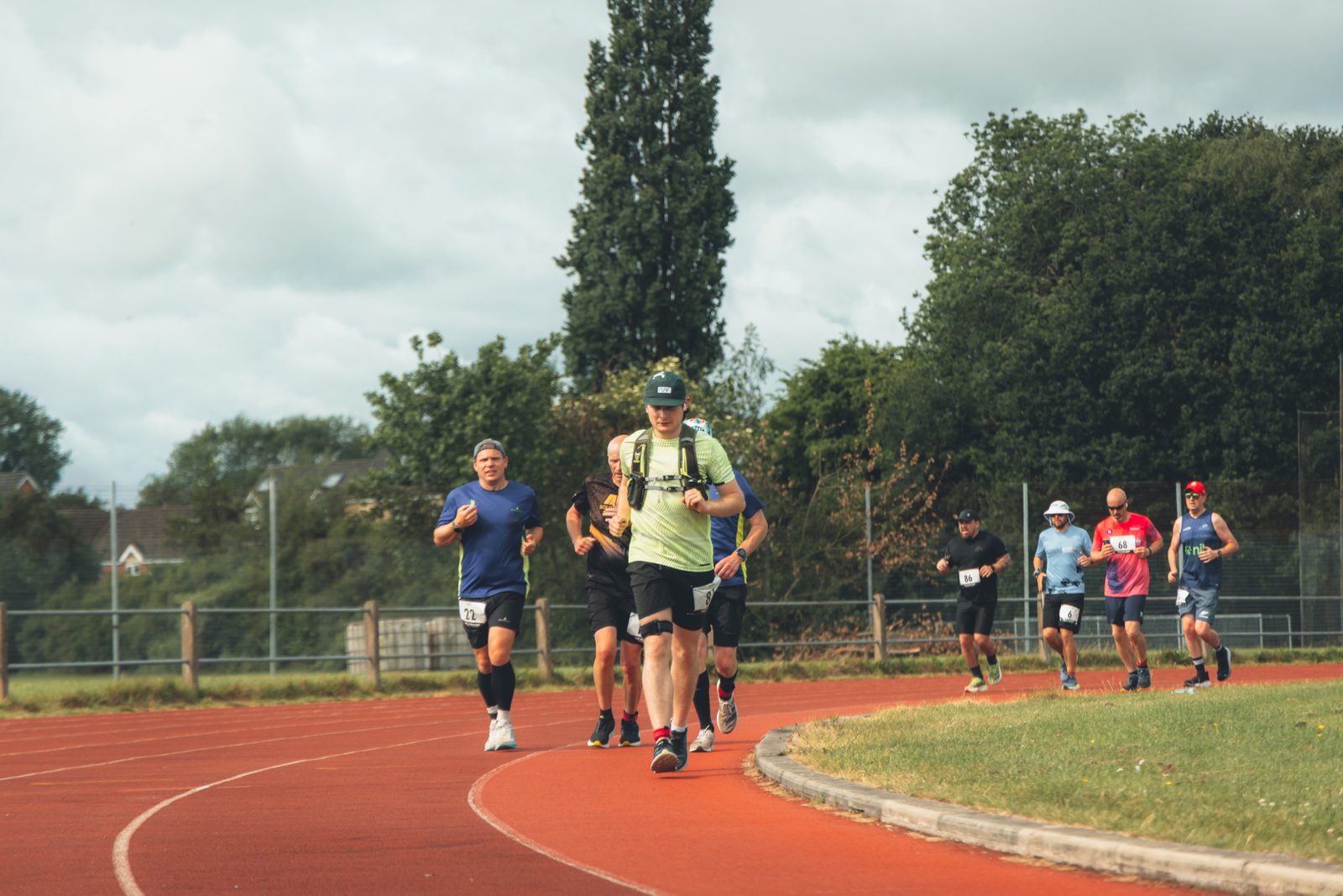 A group of runners, each wearing athletic gear and numbered bibs, run on a red track surrounded by greenery and trees on a cloudy day. The lead runner wears a light-yellow shirt and a cap.