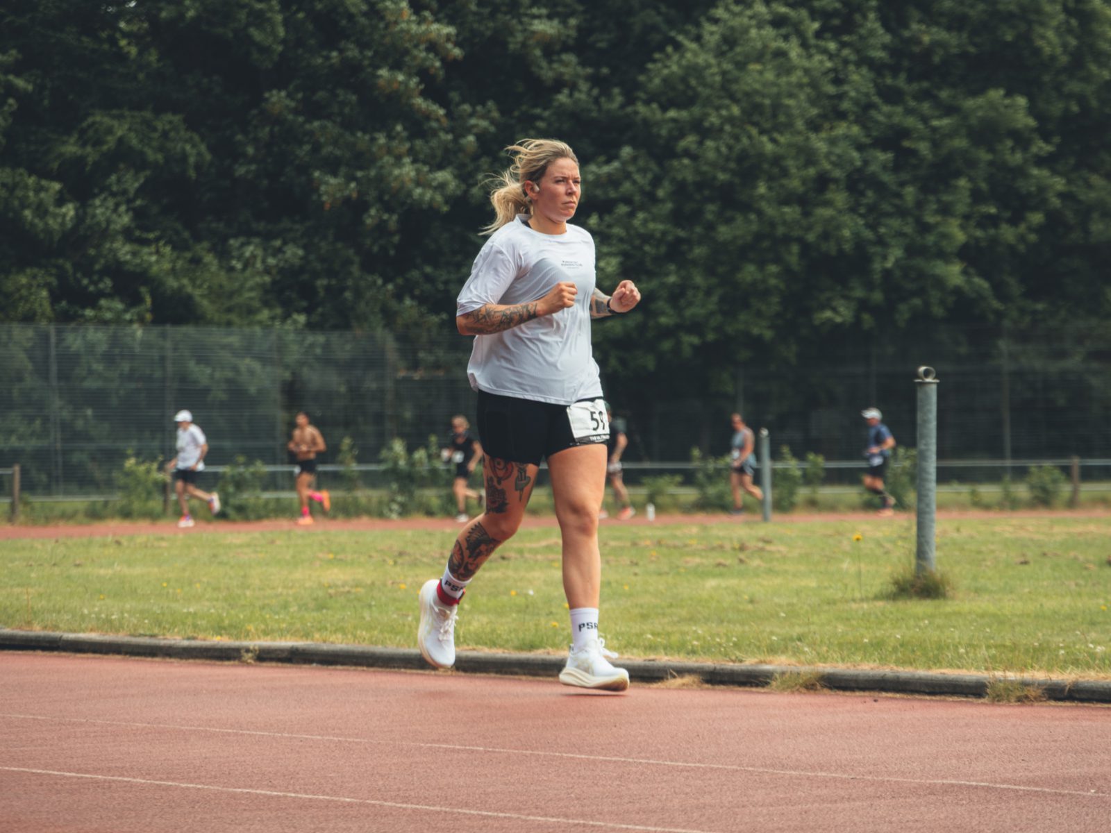 A woman with tattoos jogs on an outdoor track, wearing a white shirt, shorts, and running shoes. There are people and trees in the background.