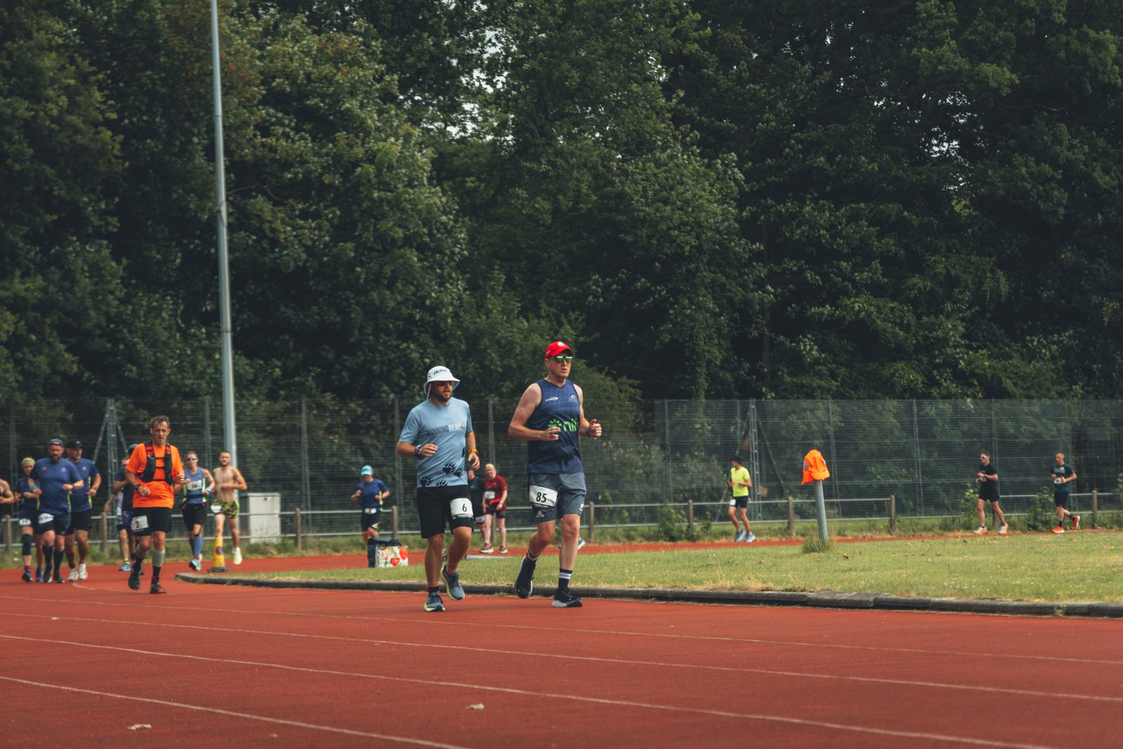 Two men run on a red track surrounded by trees, with other runners and people in the background. Both wear race bibs; one wears a white hat, the other a red cap. The scene appears to be part of a race or marathon event.