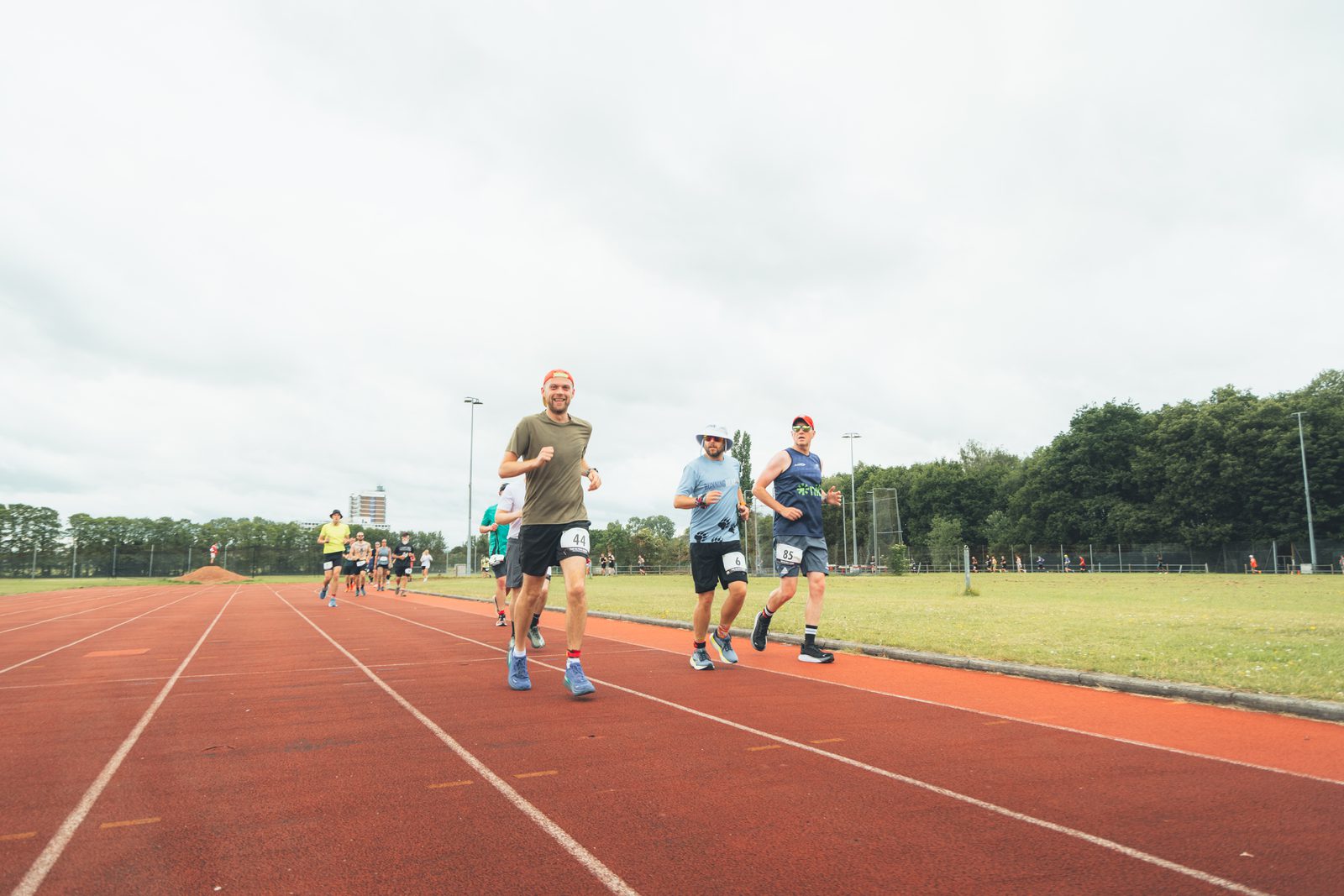 A group of runners participate in a race on an outdoor track, with three men in the foreground smiling as they run. Other runners follow behind. The sky is overcast and trees surround the track.