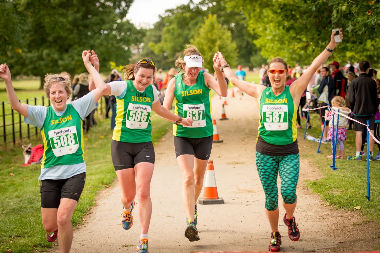 Four jubilant runners wearing matching green and yellow "Silson" tops celebrate as they cross a finish line, holding hands and raising their arms in triumph. They wear race bibs numbered 508, 506, 587, and 531. Spectators and trees line the path, creating a vibrant atmosphere.