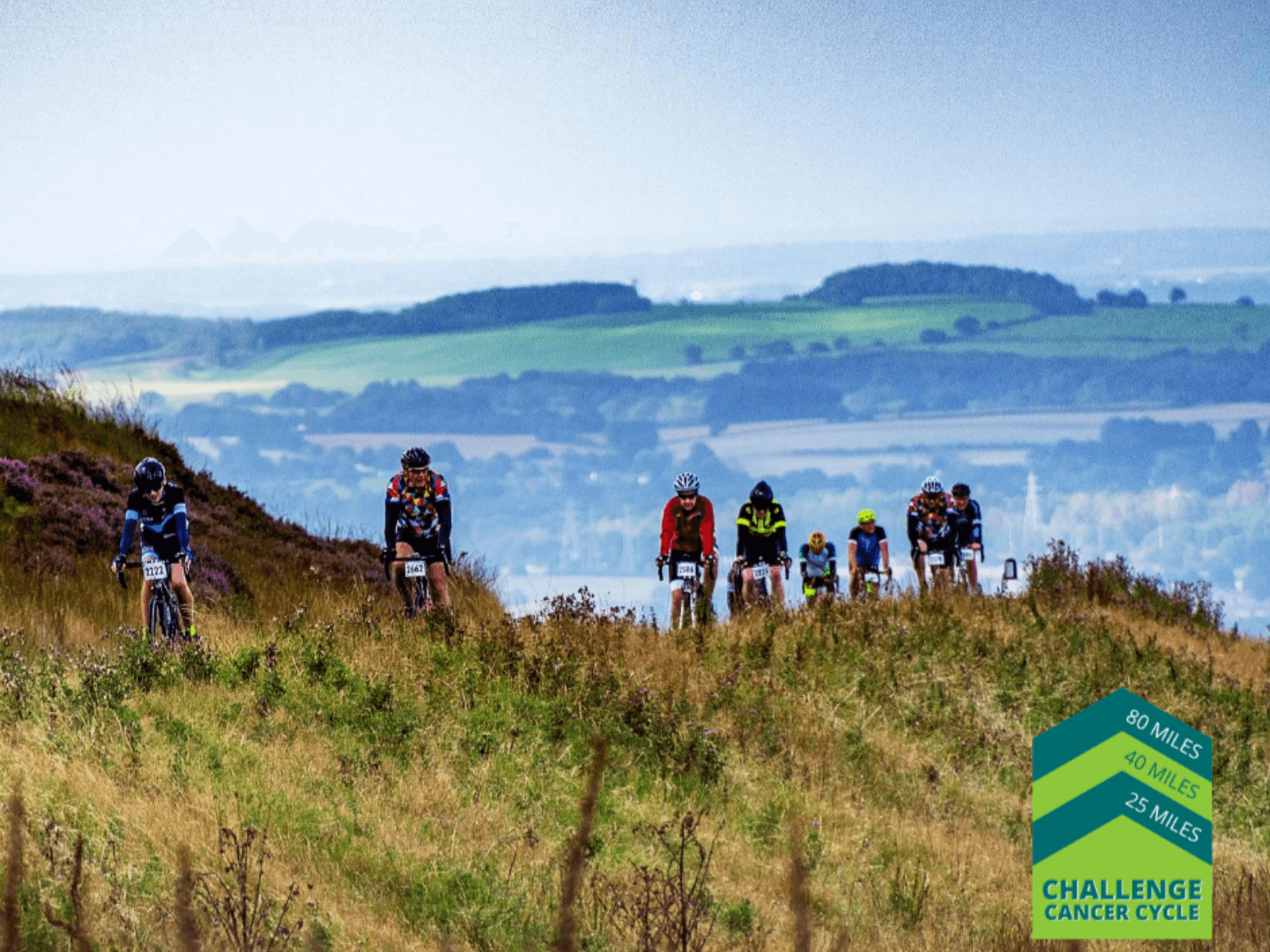 A group of cyclists rides on a grassy hillside during a cycling event. The landscape features rolling hills, forests, and fields under a clear sky. A logo in the corner reads "Challenge Cancer Cycle: 80 miles, 40 miles, 25 miles.