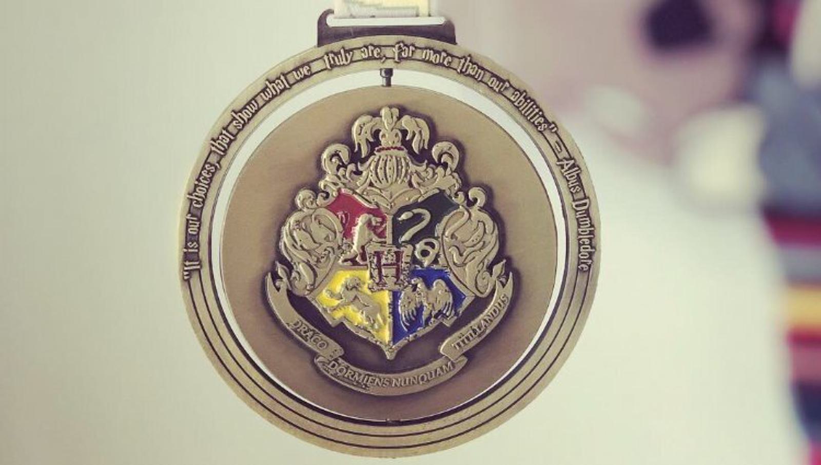 A close-up of a gold pendant featuring the Hogwarts crest from the Harry Potter series. The crest is divided into four sections, each representing one of the Hogwarts houses: Gryffindor, Hufflepuff, Ravenclaw, and Slytherin. A quote encircles the crest.