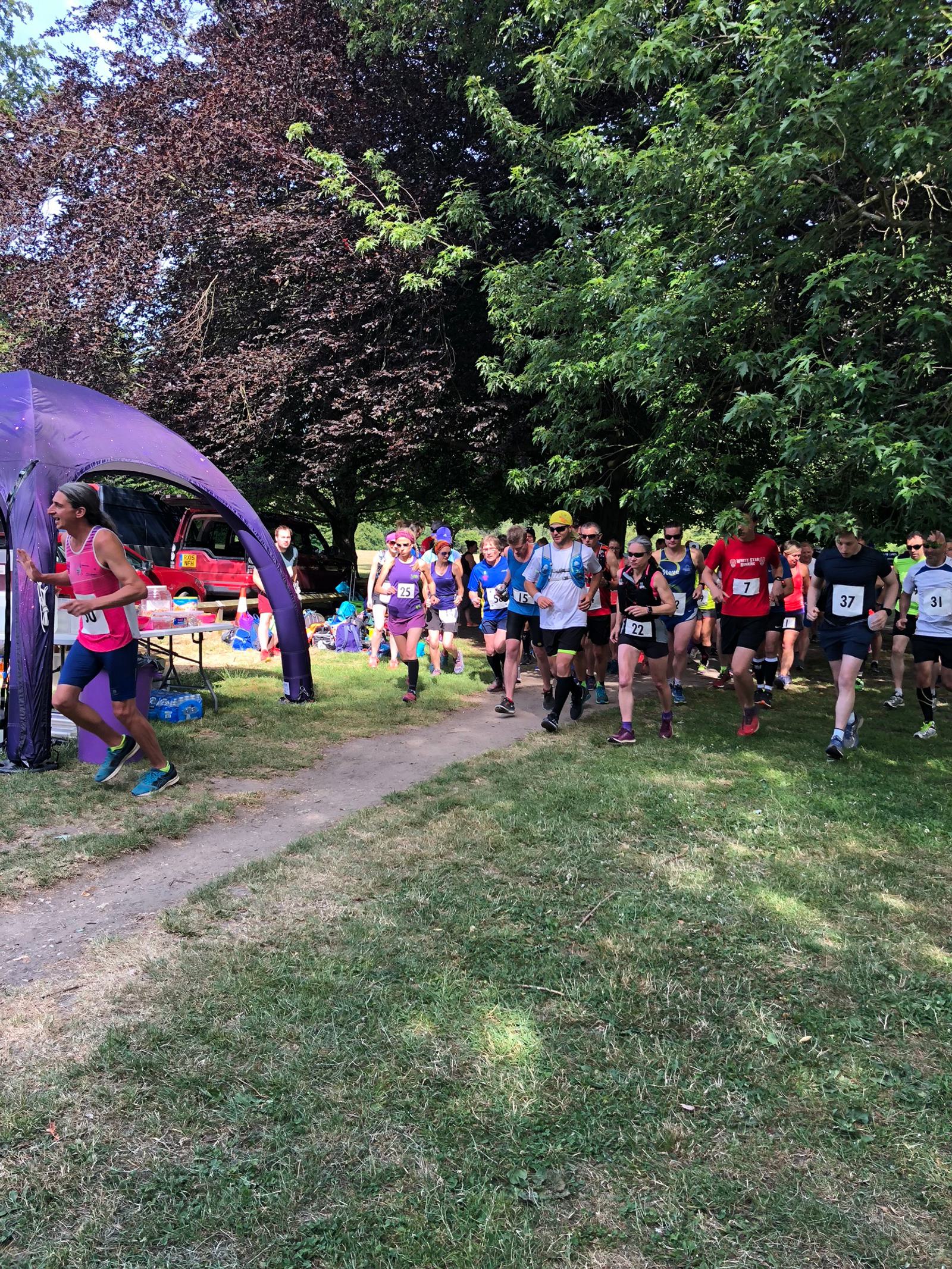 A group of runners participating in a race are seen running through a park. They wear race bibs with numbers and brightly colored athletic attire. Trees and grass surround them, and a purple canopy tent is set up nearby with people watching and cheering.