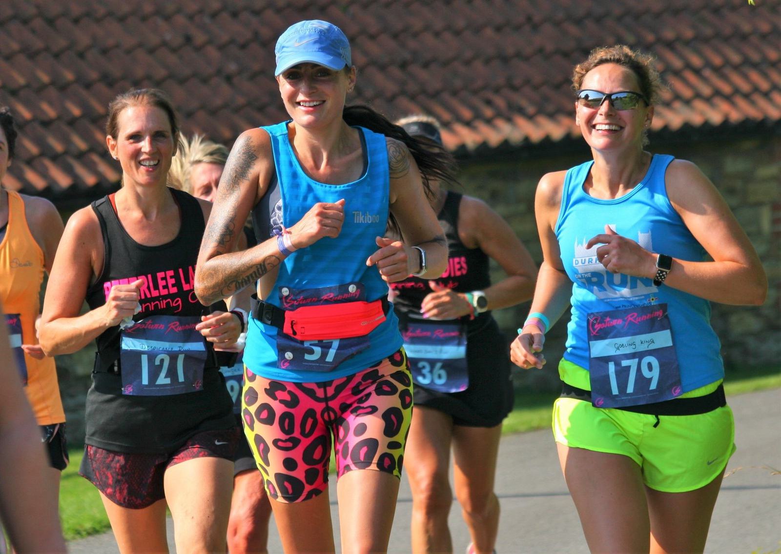 A group of runners, wearing colorful athletic gear and race bibs, smile and jog together during a sunny outdoor race. They appear joyful and energetic, with a building featuring a tiled roof visible in the background.