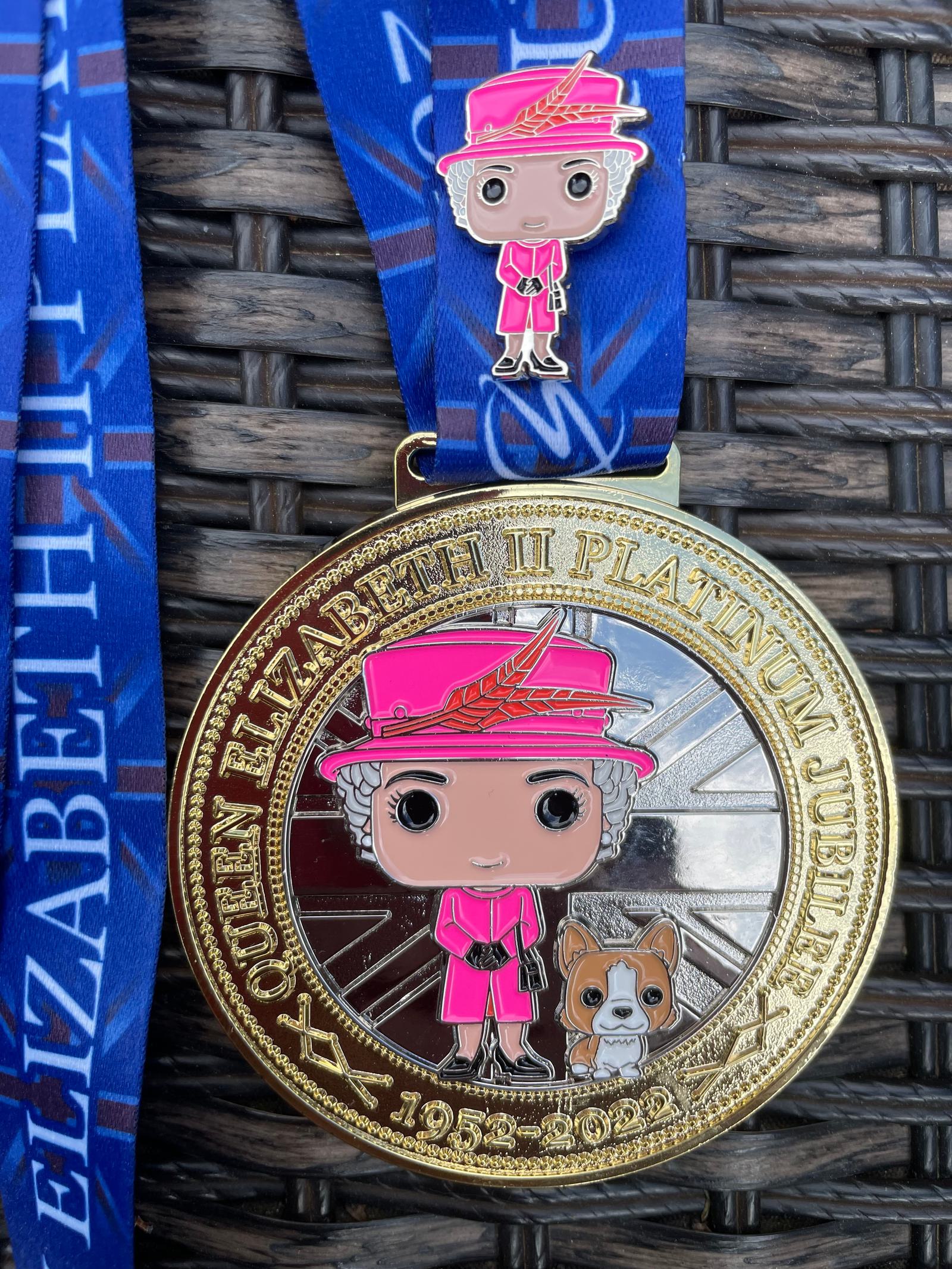 A gold medal commemorating Queen Elizabeth II's Platinum Jubilee is shown with an illustration of the Queen in a pink outfit, holding a dog. The medal has "QUEEN ELIZABETH II PLATINUM JUBILEE 1952-2022" inscribed. It hangs on a blue ribbon featuring the same text.
