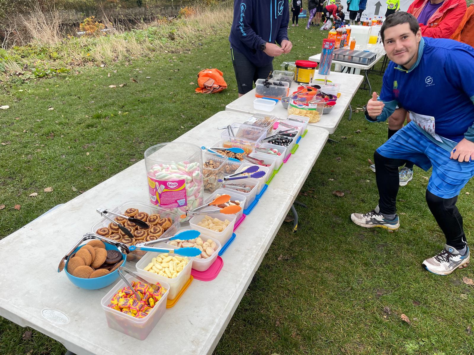 A table filled with various colorful snacks and treats, including gummy candies, cookies, chocolate pieces, marshmallows, and fruit, is displayed outdoors. A person in athletic attire stands beside the table, giving a thumbs-up gesture and smiling at the camera.