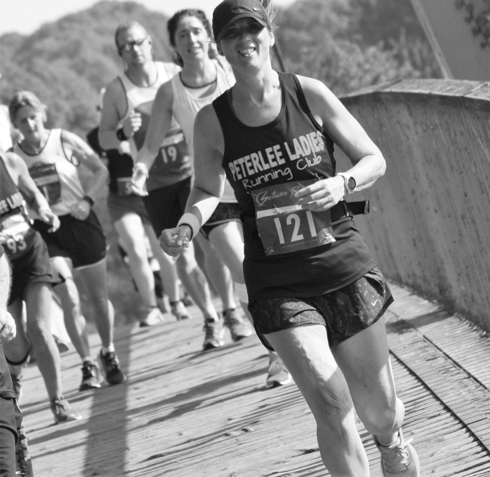 Black and white photo of a group of runners participating in a race on a bridge. A smiling woman in the foreground is wearing a cap, sleeveless top that says "Peterlee Ladies Running Club," and a bib with the number 121. Other runners are visible in the background.
