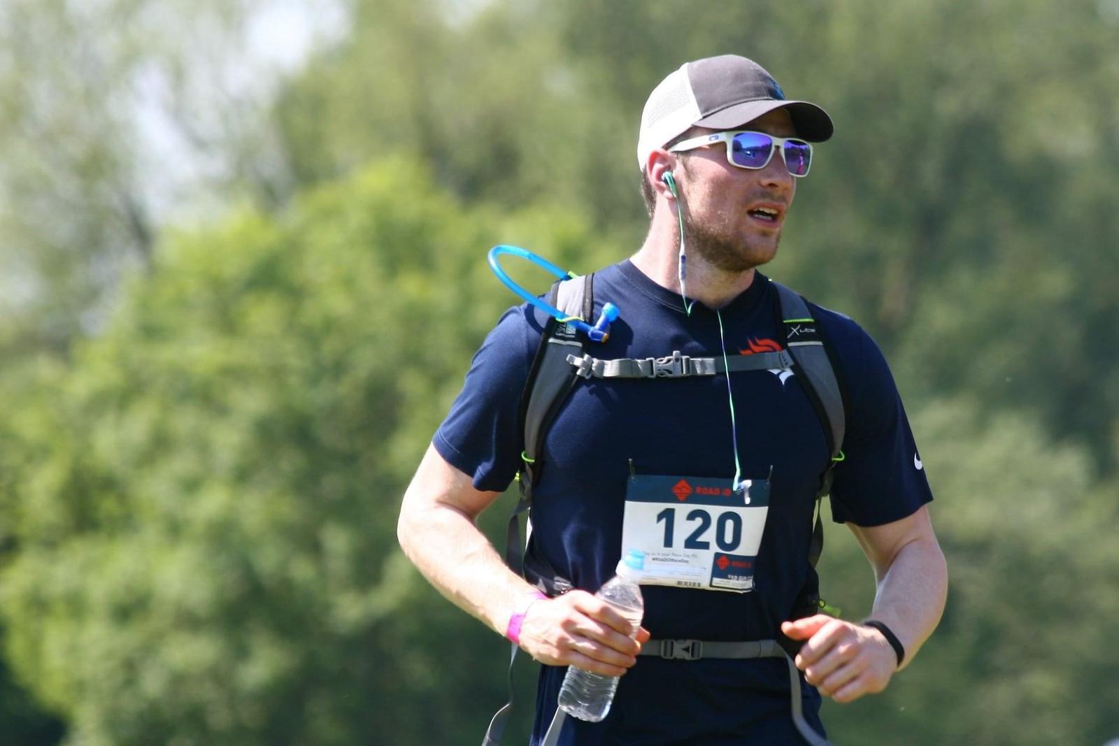 A man participating in an outdoor race, wearing a black cap and sunglasses. He is dressed in a navy blue shirt and has a hydration pack on his back. He is holding a water bottle and his race bib number is 120. Lush green trees are visible in the background.