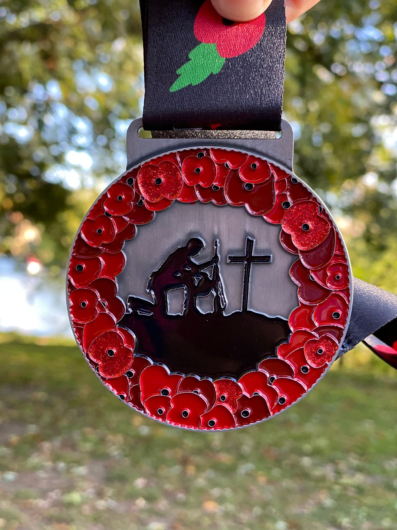 A round medal features a silhouette of a soldier kneeling before a cross, with a rifle at his side. The medal is decorated with a border of vibrant red poppies. Blurred foliage and sky create a natural background. The medal hangs from a black ribbon.