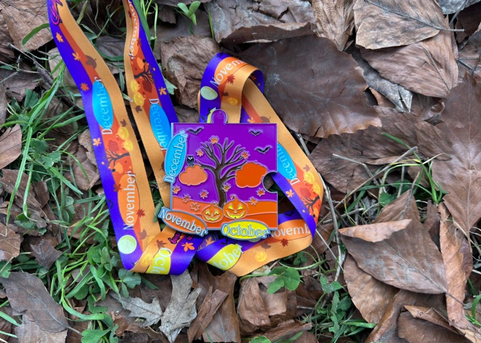 A colorful medal with a Halloween theme featuring a tree, pumpkins, and bats. The ribbon attached has autumnal colors and displays the months "October," "November," and "December." The medal is placed on fallen leaves and grass.