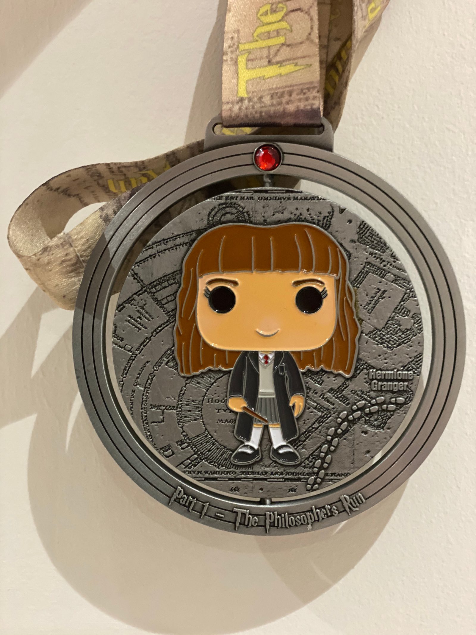 A medal featuring a cartoonish figure of a girl with brown hair in a school uniform. The background has an intricate design, and the medal reads, "Part 1 - The Philosopher's Run." The ribbon attached to the medal is light-colored with text printed on it.