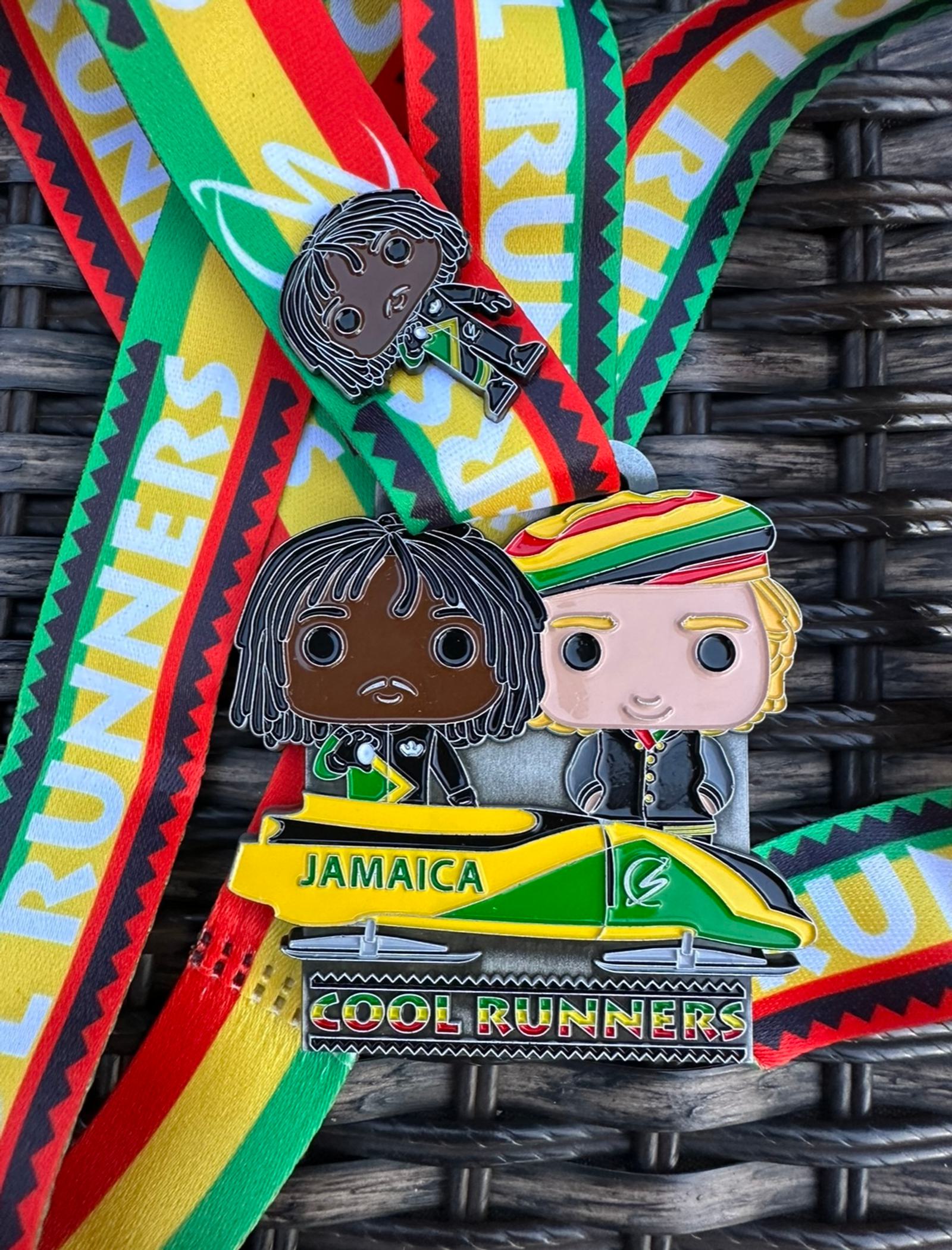 Close-up of two Jamaica-themed medals against a braided background. One medal features two cartoon characters in a bobsled and reads "Jamaica" and "Cool Runners." The other has a single cartoon character. Both medals are attached to colorful ribbons.