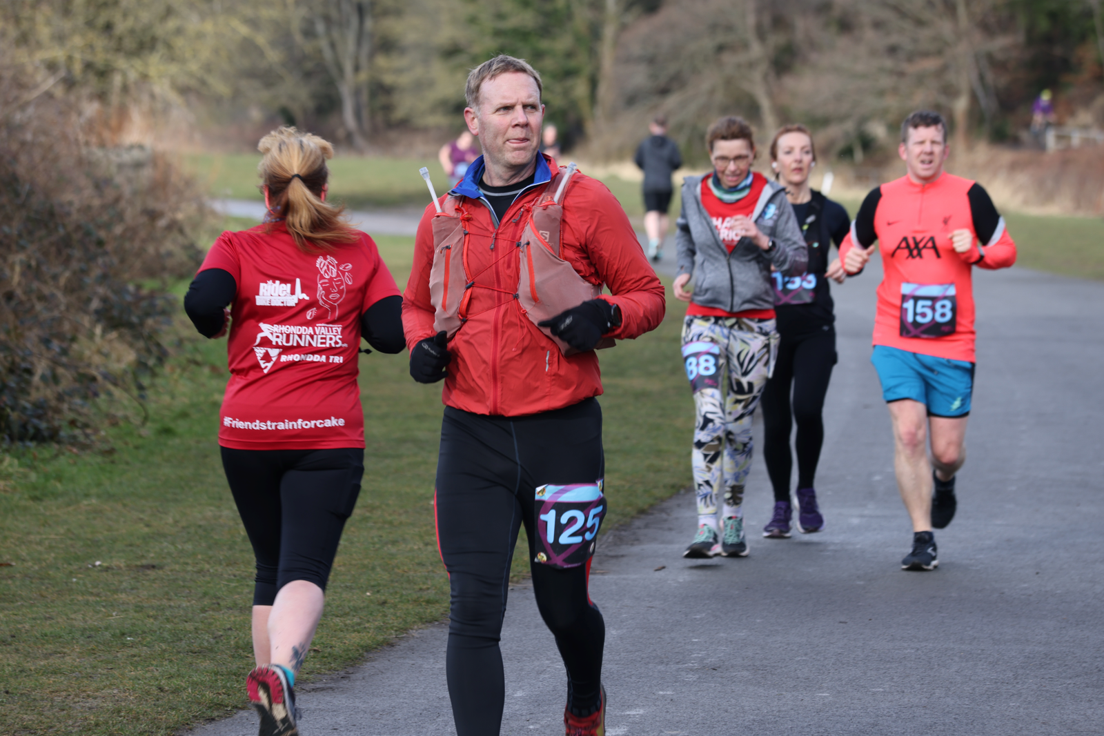 A group of runners participating in an outdoor race on a paved path in a wooded park. The runners are wearing race bibs, sporty attire, and some have hydration packs. One runner, wearing a red jacket and black pants, is in the forefront with bib number 125.