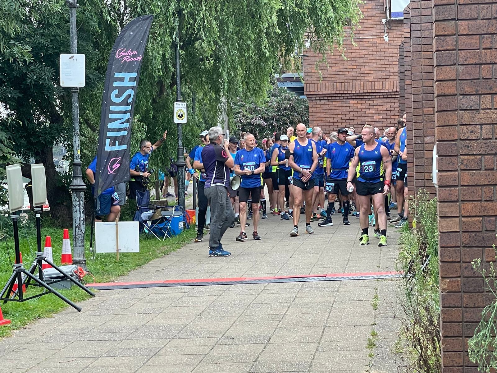 Runners dressed in blue shirts gather at the finish line of a race. A man in a grey shirt is holding a megaphone, giving instructions. A large black flag with "FINISH" in white letters is displayed. Trees and a brick building are visible in the background.