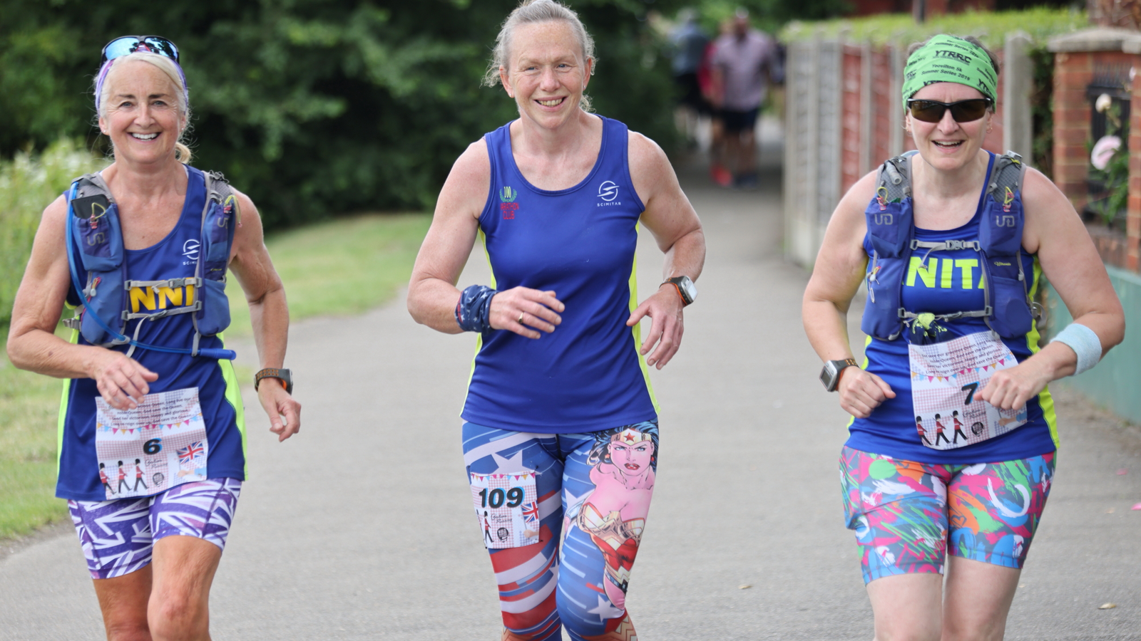 Three women smiling and jogging outside on a paved path, wearing colorful athletic clothes and race bibs. Trees, grass, flowers, and brick walls are in the background. All three appear happy and energetic.