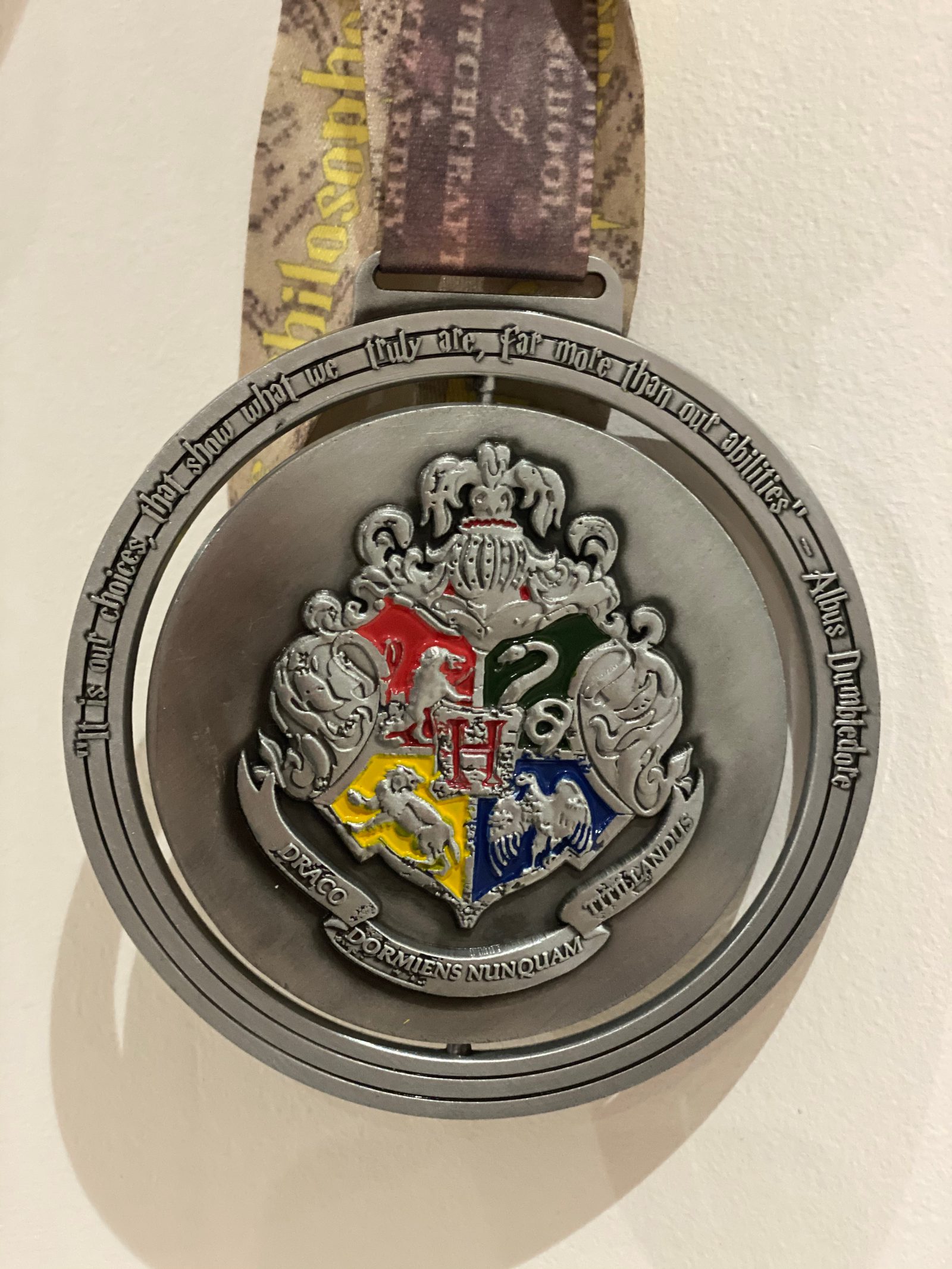 A close-up image of a gray, metallic Harry Potter-themed medal. The medal features the Hogwarts crest with four quadrants representing the houses: Gryffindor, Slytherin, Hufflepuff, and Ravenclaw. The ribbon attached has a pattern with legible text.