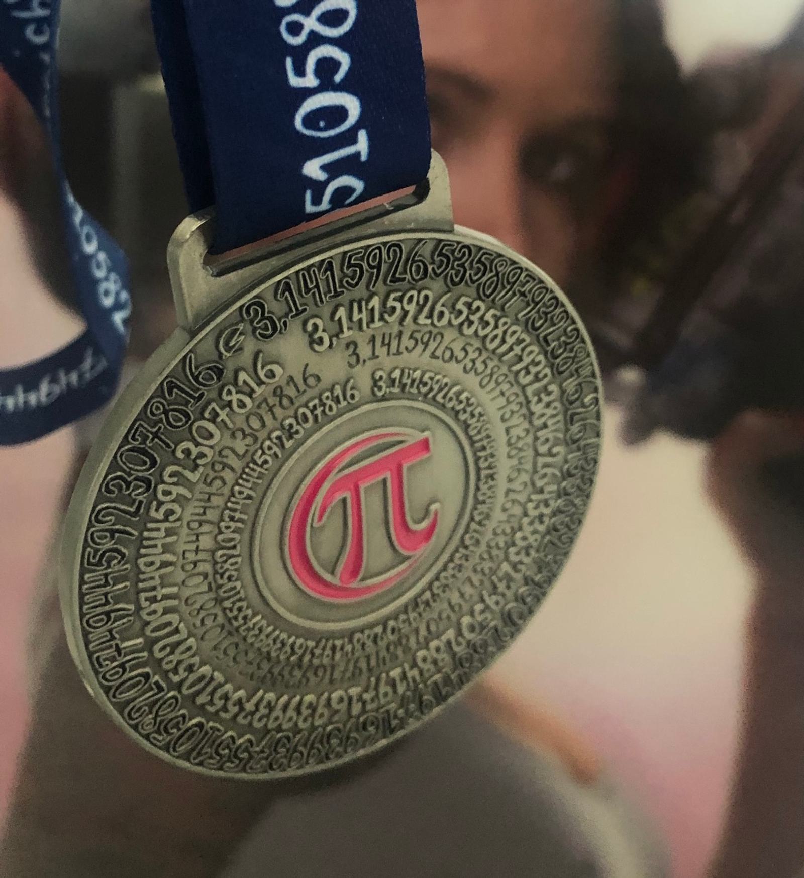 A close-up of a Pi Day medal with the Pi symbol in the center, surrounded by the digits of Pi engraved in a spiral. The medal is attached to a blue ribbon with white text. A blurred background features the out-of-focus face of a person.