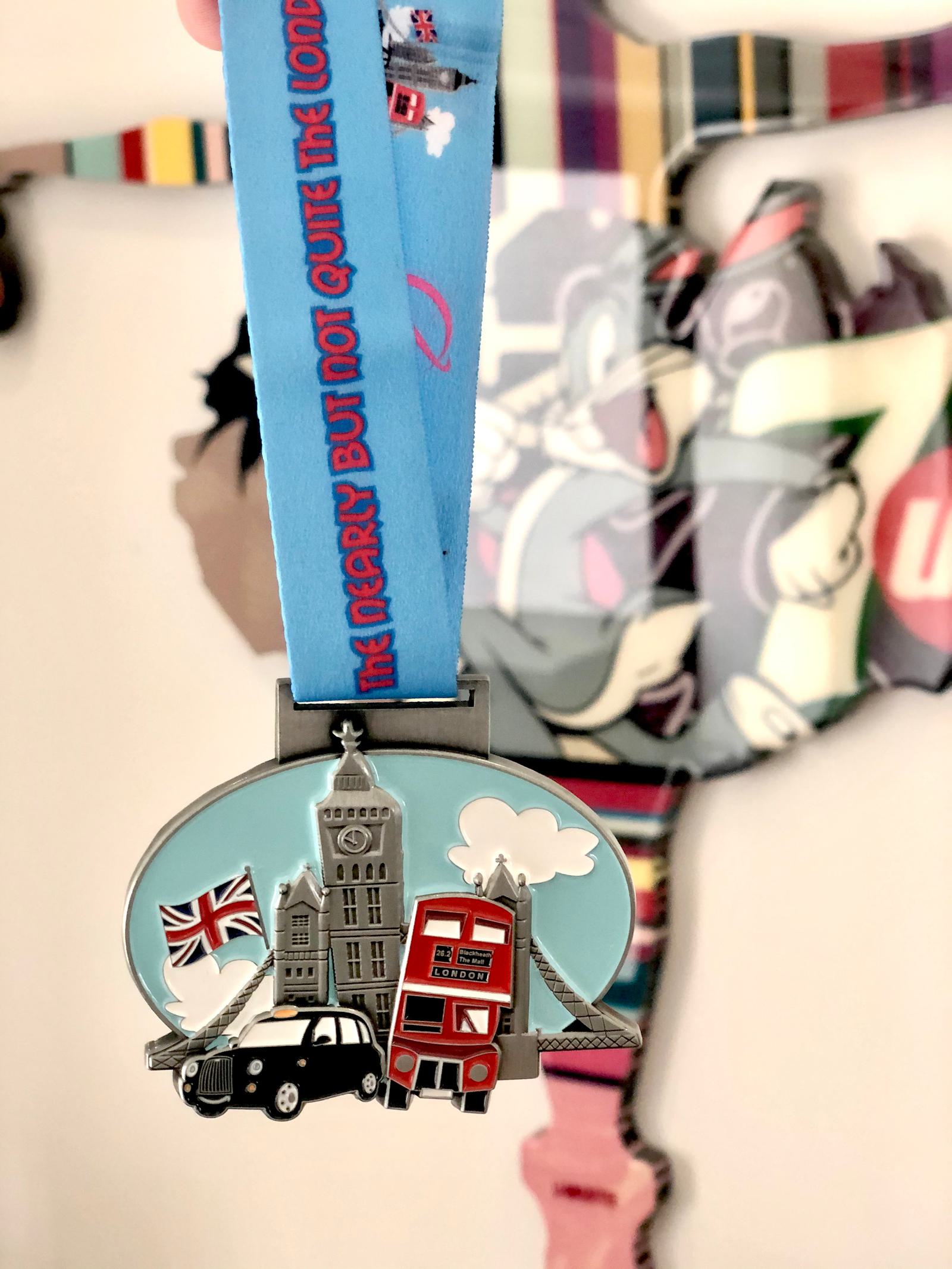 A medal hangs from a blue ribbon featuring the text, "The Merry But Not Quite The London." The medal showcases iconic London symbols: Big Ben, a red double-decker bus, a black cab, and the London Eye. A colorful wall art background is partially visible.