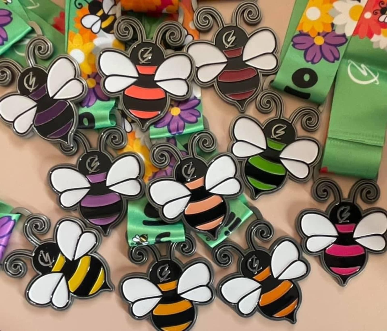 A collection of bee-themed medals with striped bodies in various colors (red, orange, yellow, green, pink, purple), and green lanyards featuring floral patterns and symbols. The medals have large wings and curly antennae.
