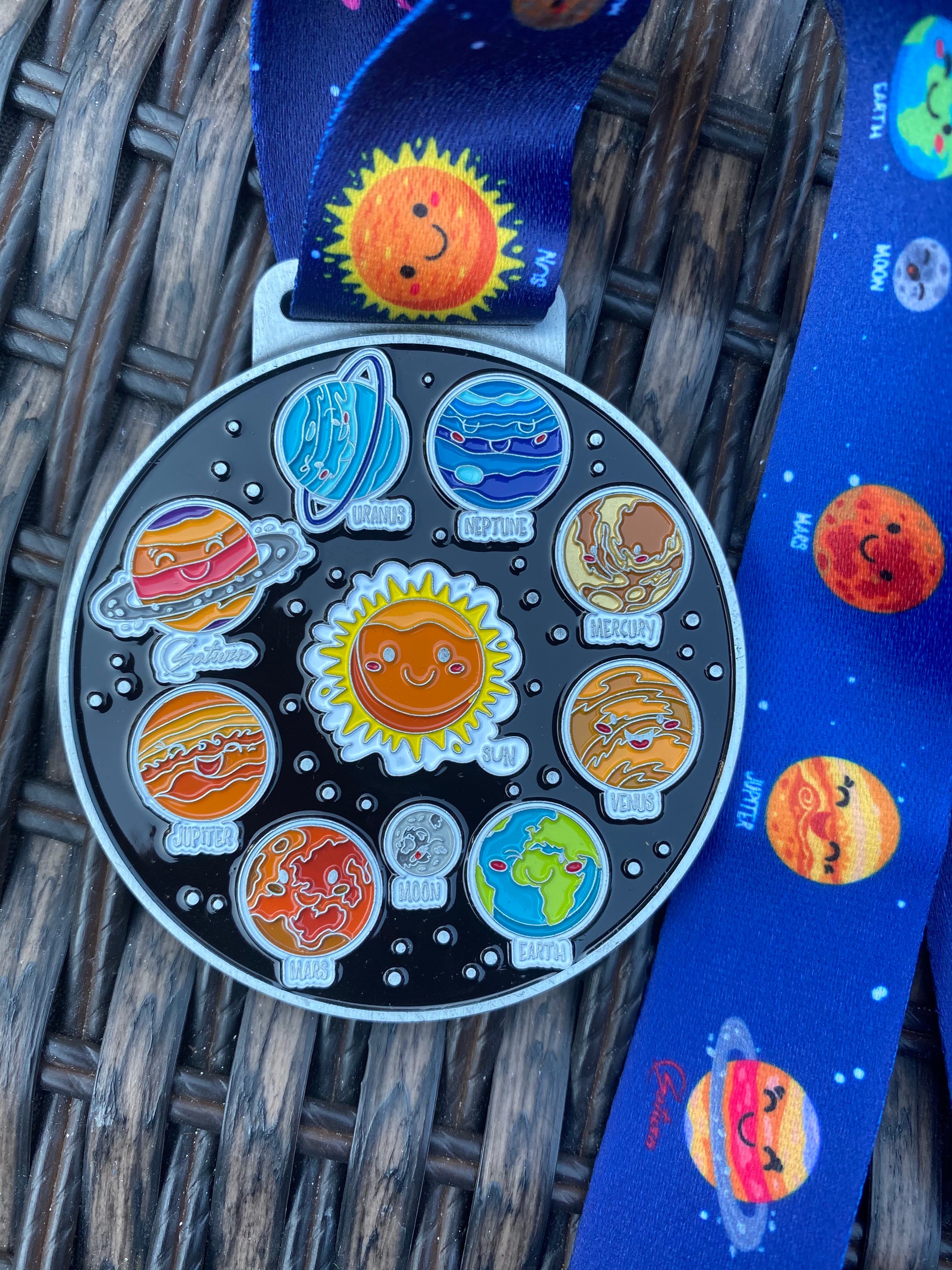 A colorful medal with cartoon representations of the planets in the solar system arranged around the sun. The medal hangs from a blue lanyard decorated with cheerful illustrations of the sun and planets. The background is a woven texture.