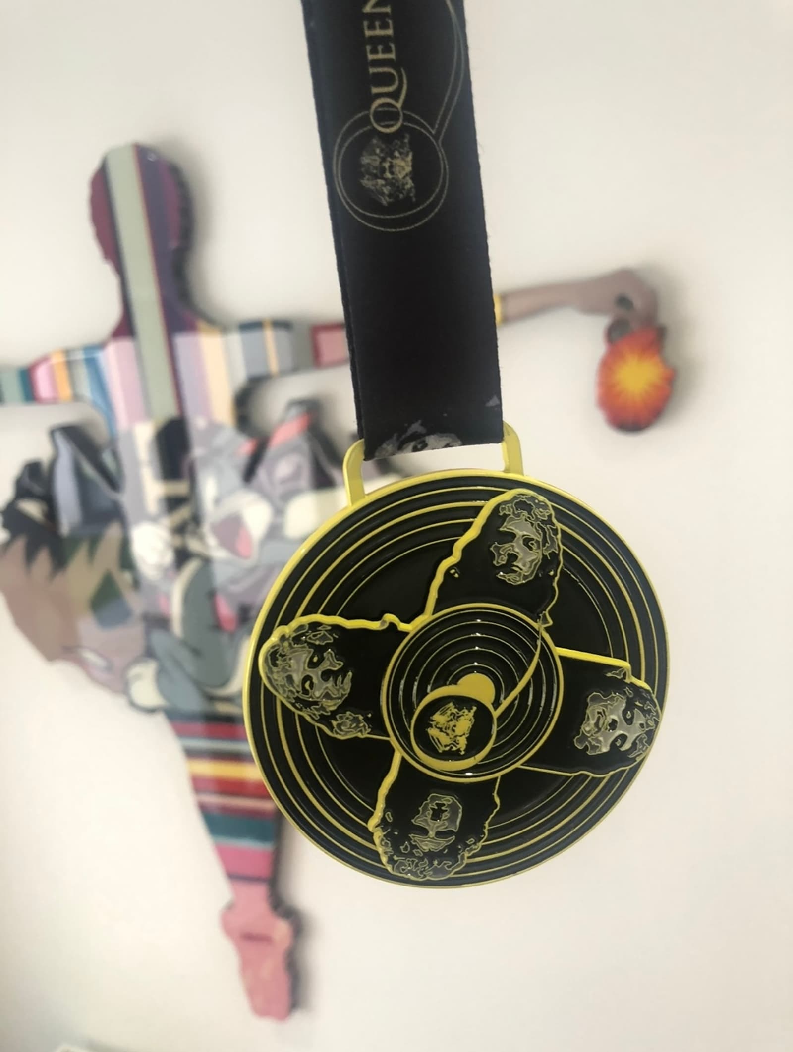 A close-up of a black and yellow circular medal with sketches of faces on it. It hangs from a black ribbon with a logo. A colorful abstract figure in the background appears blurred.