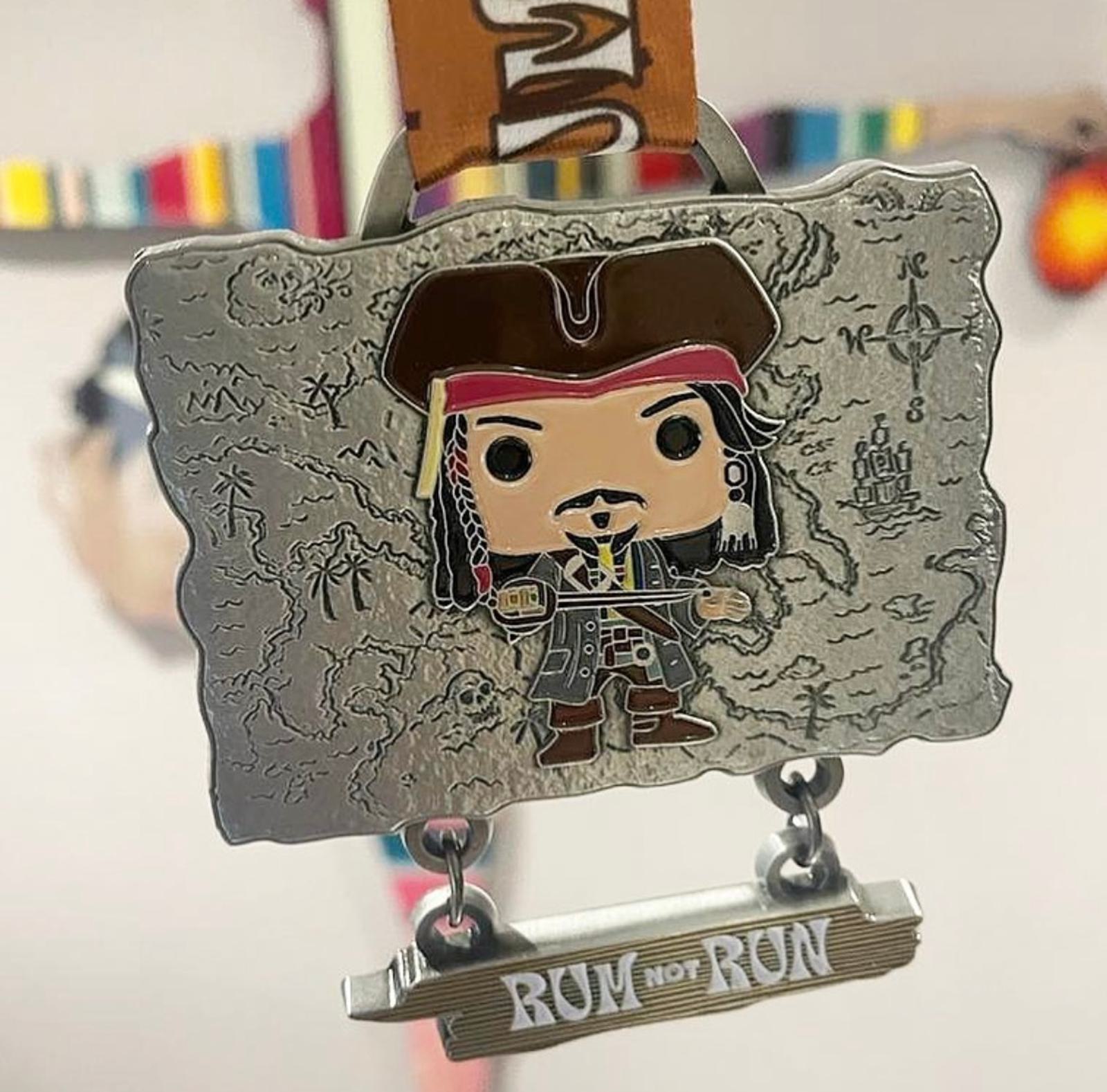 A medal designed like a treasure map featuring a cartoon pirate in the center, wearing a tricorn hat. Below the map is a rectangular bar with the phrase "RUN NOT RUN" engraved on it. The background is blurred, displaying colorful objects.