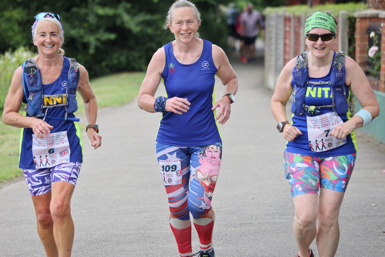 Three women participating in a race, wearing colorful athletic gear and smiling. Two are wearing blue tank tops with matching shorts featuring vibrant patterns, and one is wearing a blue tank top with knee-length shorts. They all have race bibs and hydration packs.