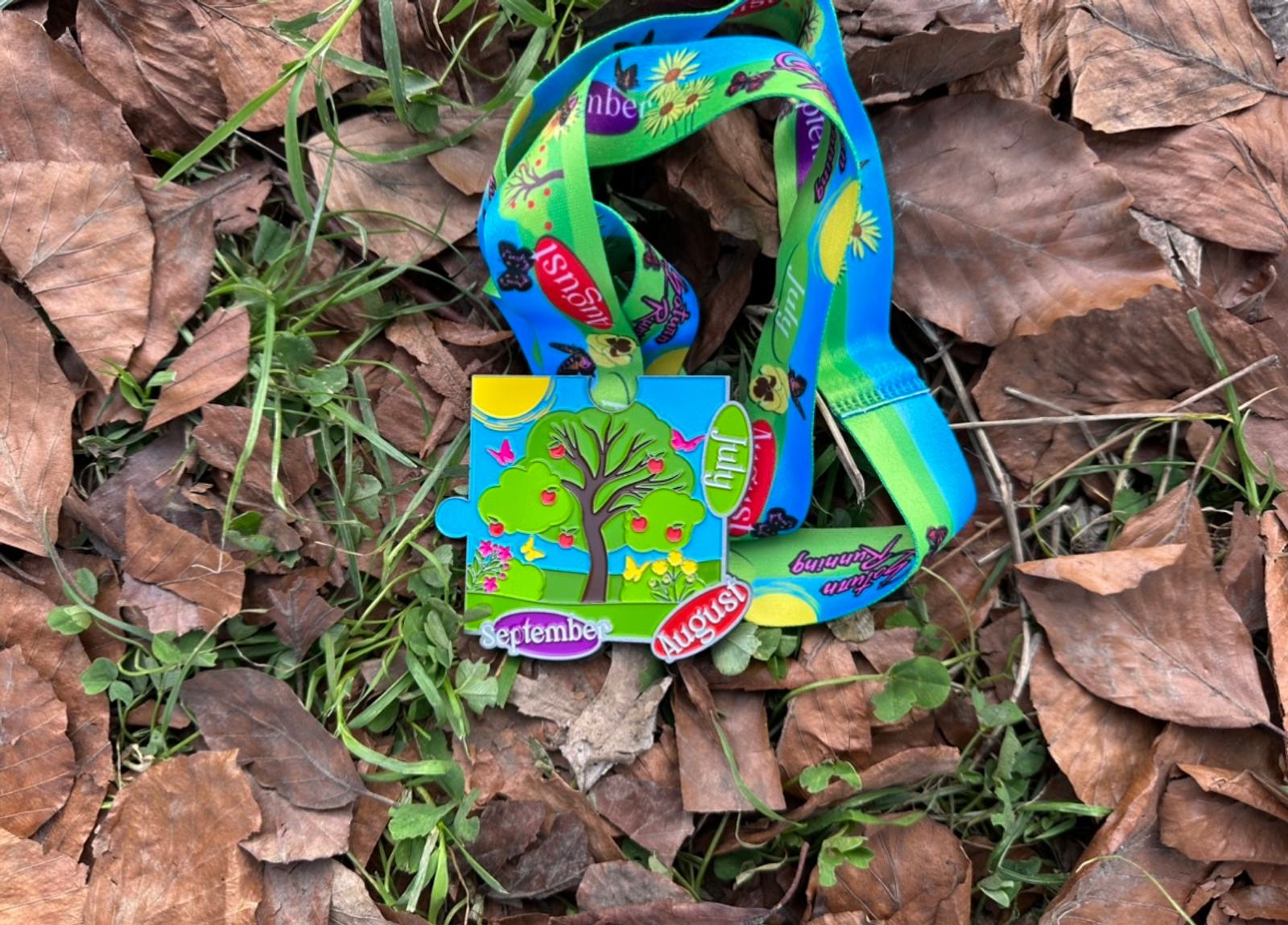 A colorful, cartoon-inspired medal with a green ribbon lies on a bed of dry brown leaves and green grass. The medal features cute green creatures, bright sun, and butterflies. The text on the ribbon reads "5 de agosto," "lamerlouvenue," and "septembre.