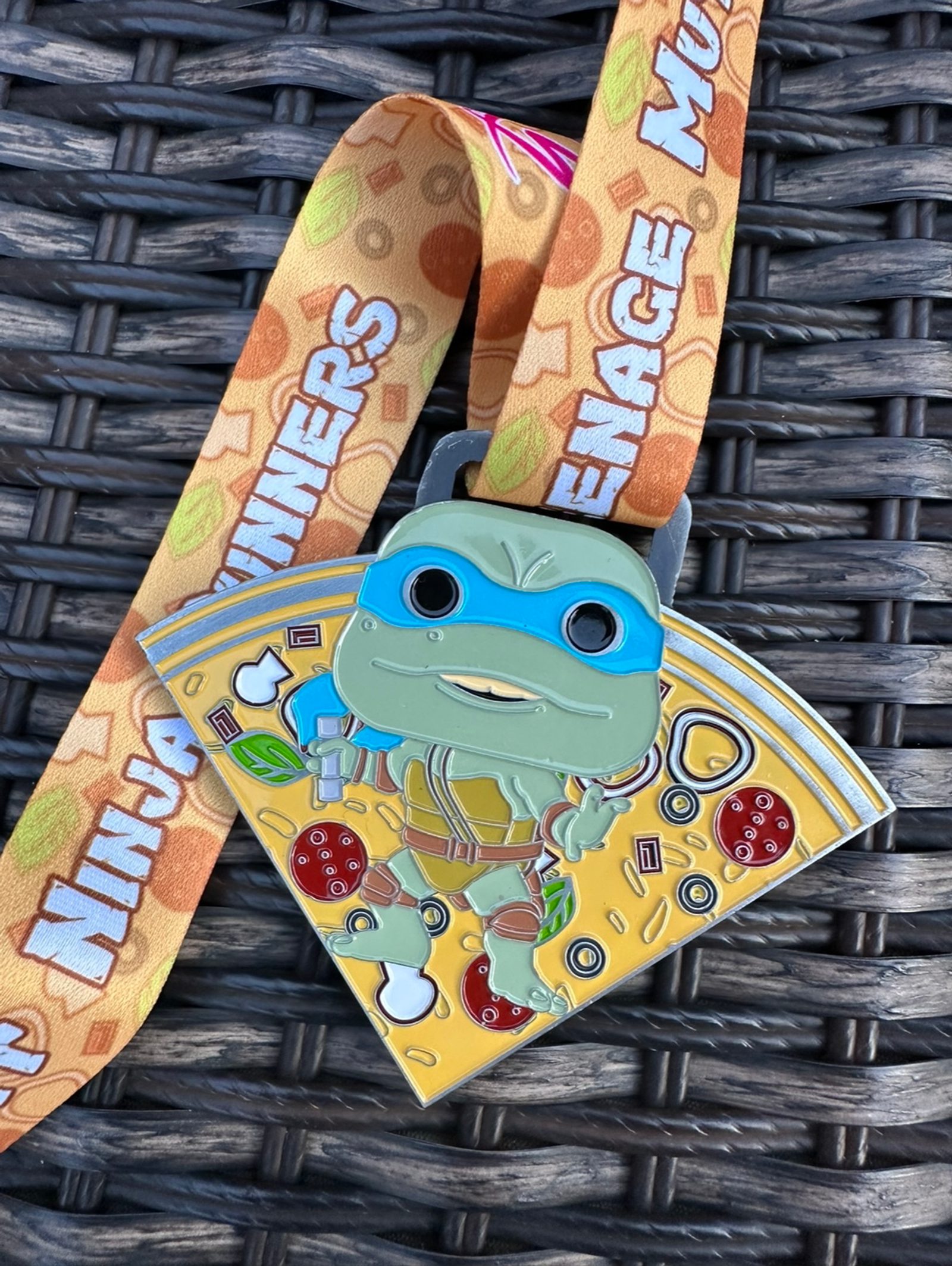 A colorful pizza-shaped medal featuring a character from Teenage Mutant Ninja Turtles. The medal shows a turtle wearing a blue mask and holding a weapon, attached to an orange lanyard with the text "Teenage Mutant Ninja Winners" printed in white.