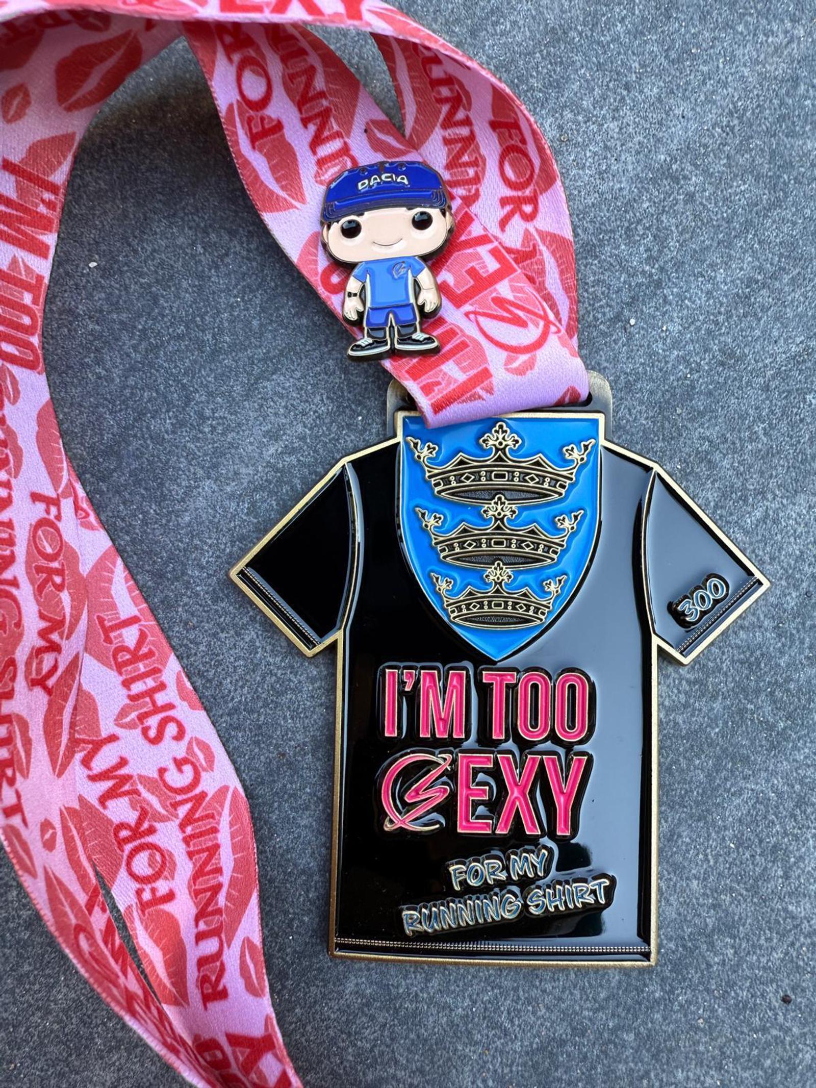 A medal with a ribbon, featuring a shirt design. The medal reads "I'm Too Sexy For My Running Shirt" in bold letters. Attached is a small figure wearing a blue cap and outfit. The ribbon is red with repeating text.