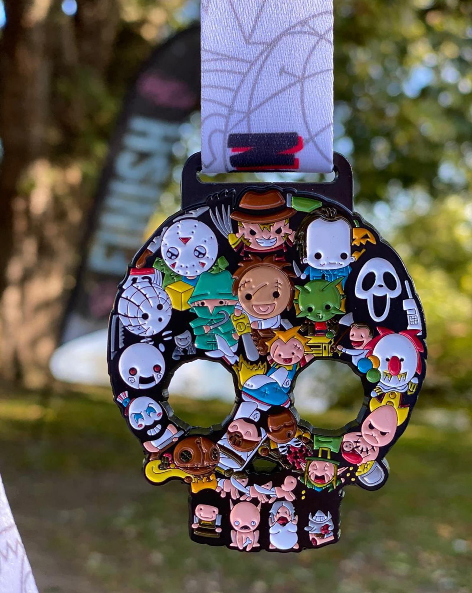 A Halloween-themed medal featuring various cartoon characters in vibrant costumes. The medal is attached to a lanyard with a blurred outdoor background of trees and a banner. The design includes elements such as a skull shape and numerous small character illustrations.