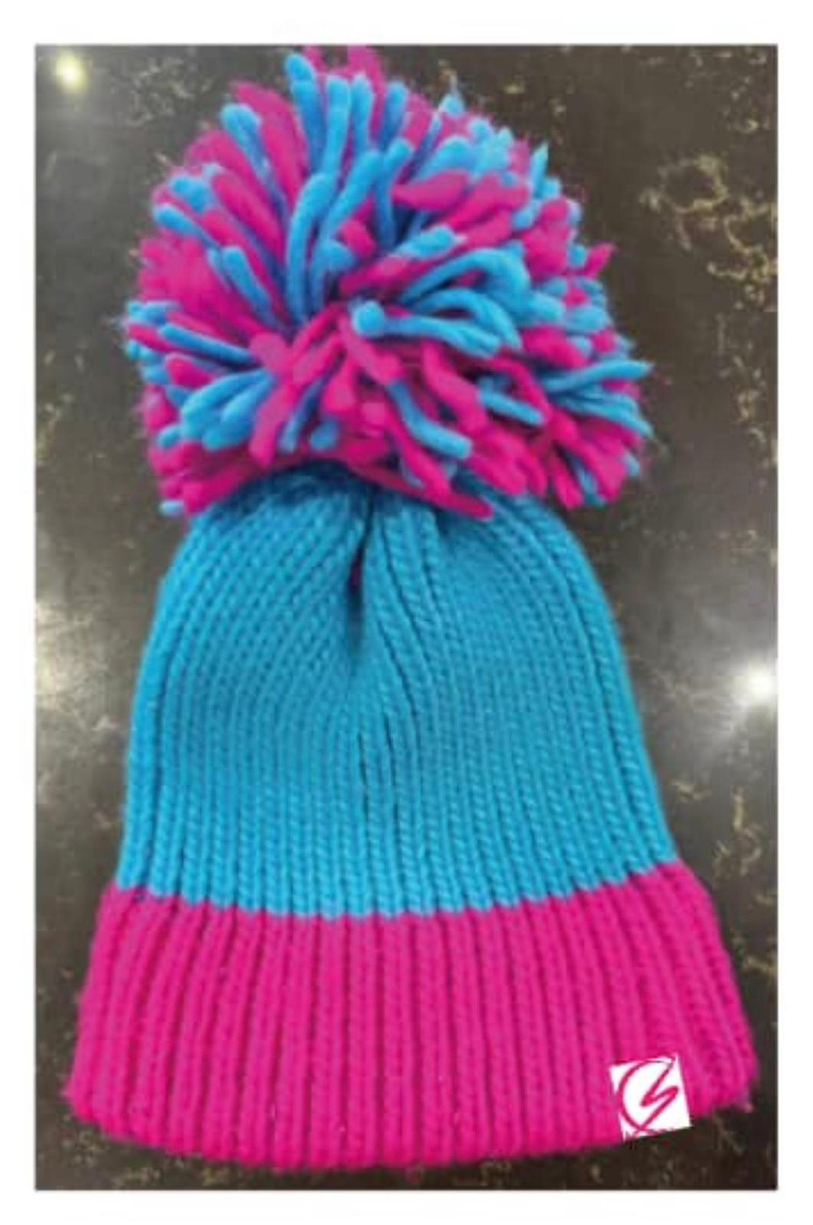 A knitted beanie with a large pom-pom on top. The lower portion of the beanie is pink, and the upper portion is blue. The pom-pom is a mix of pink and blue strands. The beanie is placed on a dark surface.