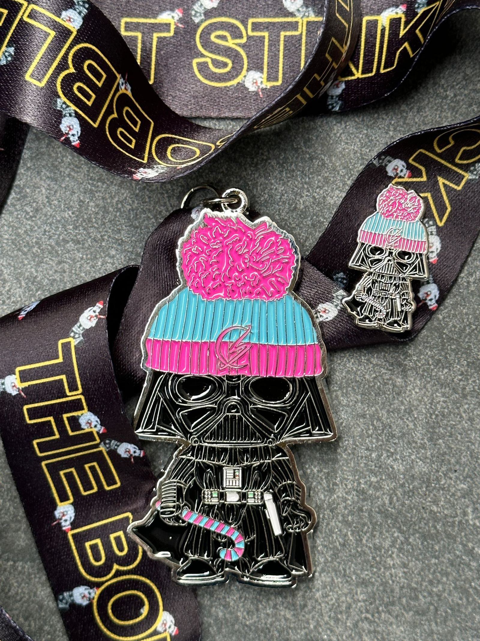 A keychain featuring a character resembling Darth Vader in a pink and blue beanie, holding a candy cane. The character is attached to a ribbon with "THE BOLT STRIKE" text and skull patterns. A smaller version of the same character is also present on the ribbon.