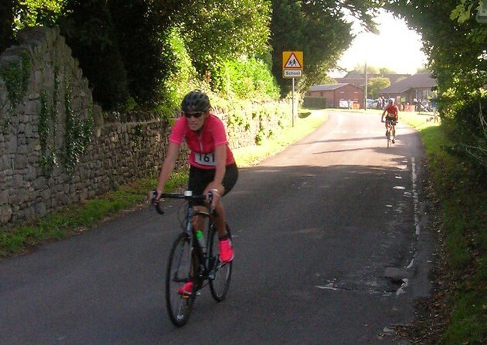 A cyclist wearing a pink shirt, helmet, and sunglasses rides on a tree-lined road near a stone wall. Another cyclist is seen in the background. A sign indicating a school is visible ahead on the right side of the road. Bright sunlight filters through the trees.