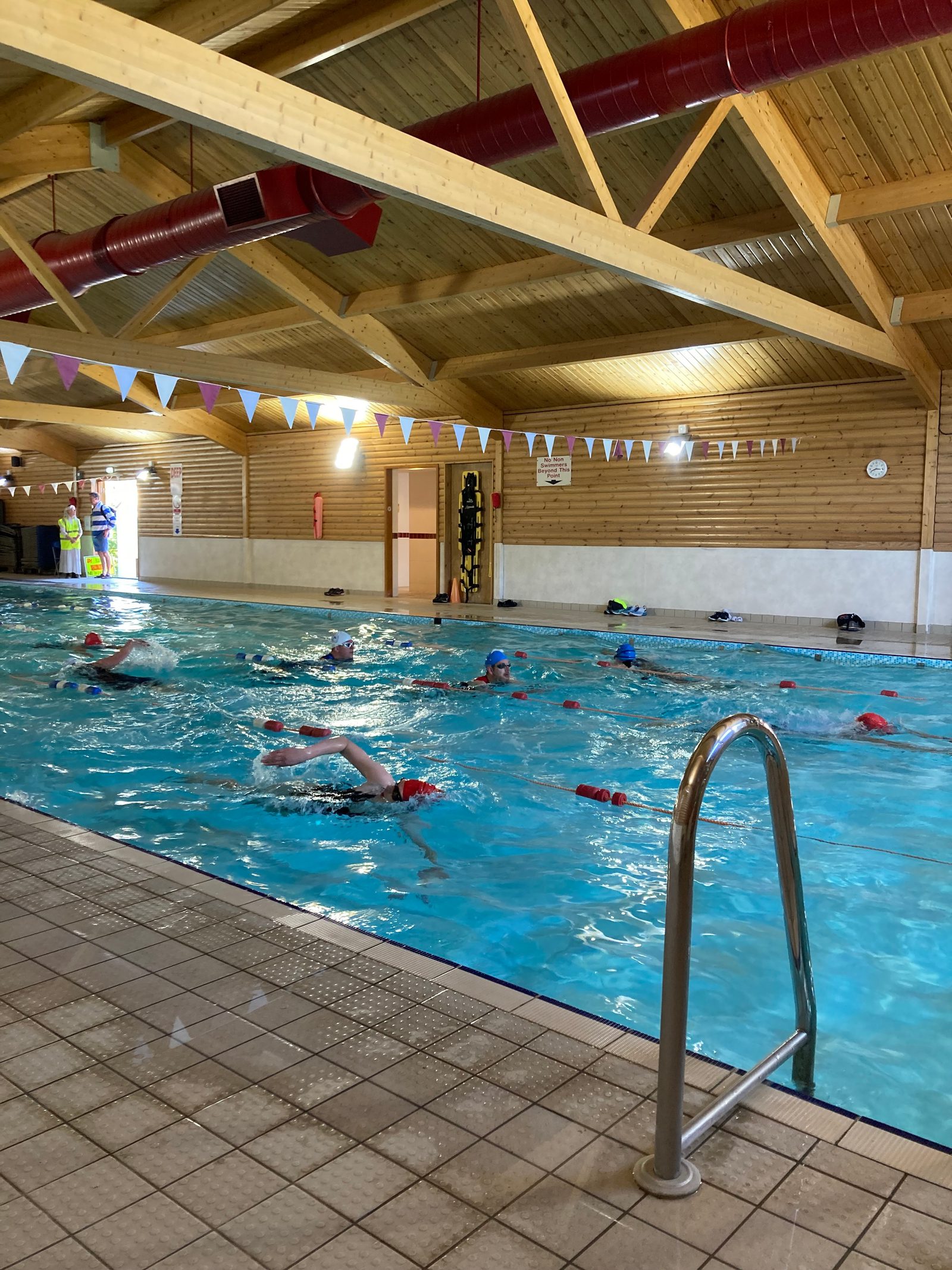 A group of people swimming in an indoor pool with wooden ceilings and walls. The pool is divided by a line of floating buoys, and a metal handrail is visible in the foreground. Decorations and a clock are on the wall in the background.