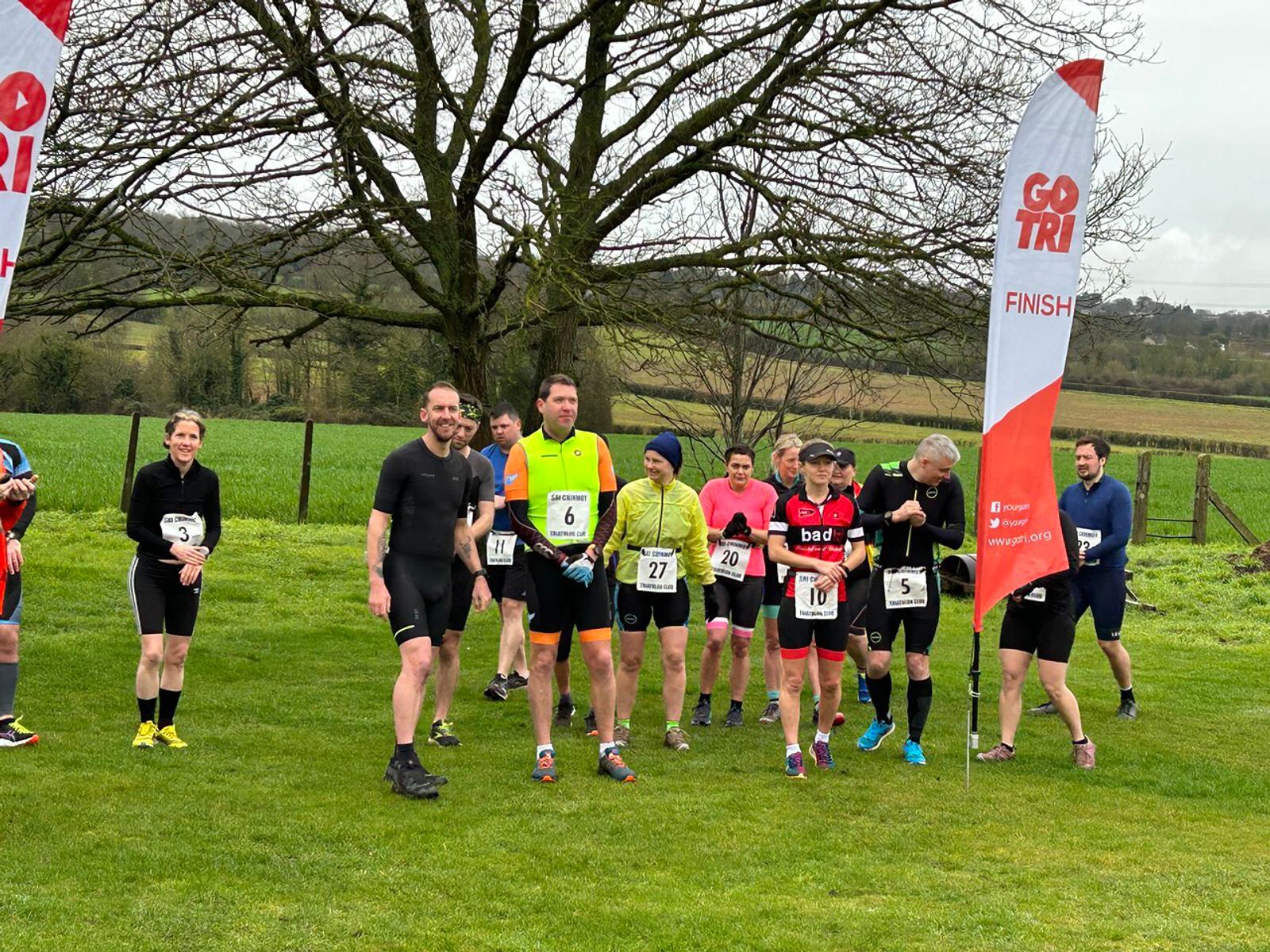 A group of participants, wearing numbers, stand at the starting line of a race on a grassy field. Some wear running gear while others are on bicycles. A "GO TRI" banner marking the finish line is visible to the right. Trees and a rural landscape are in the background.