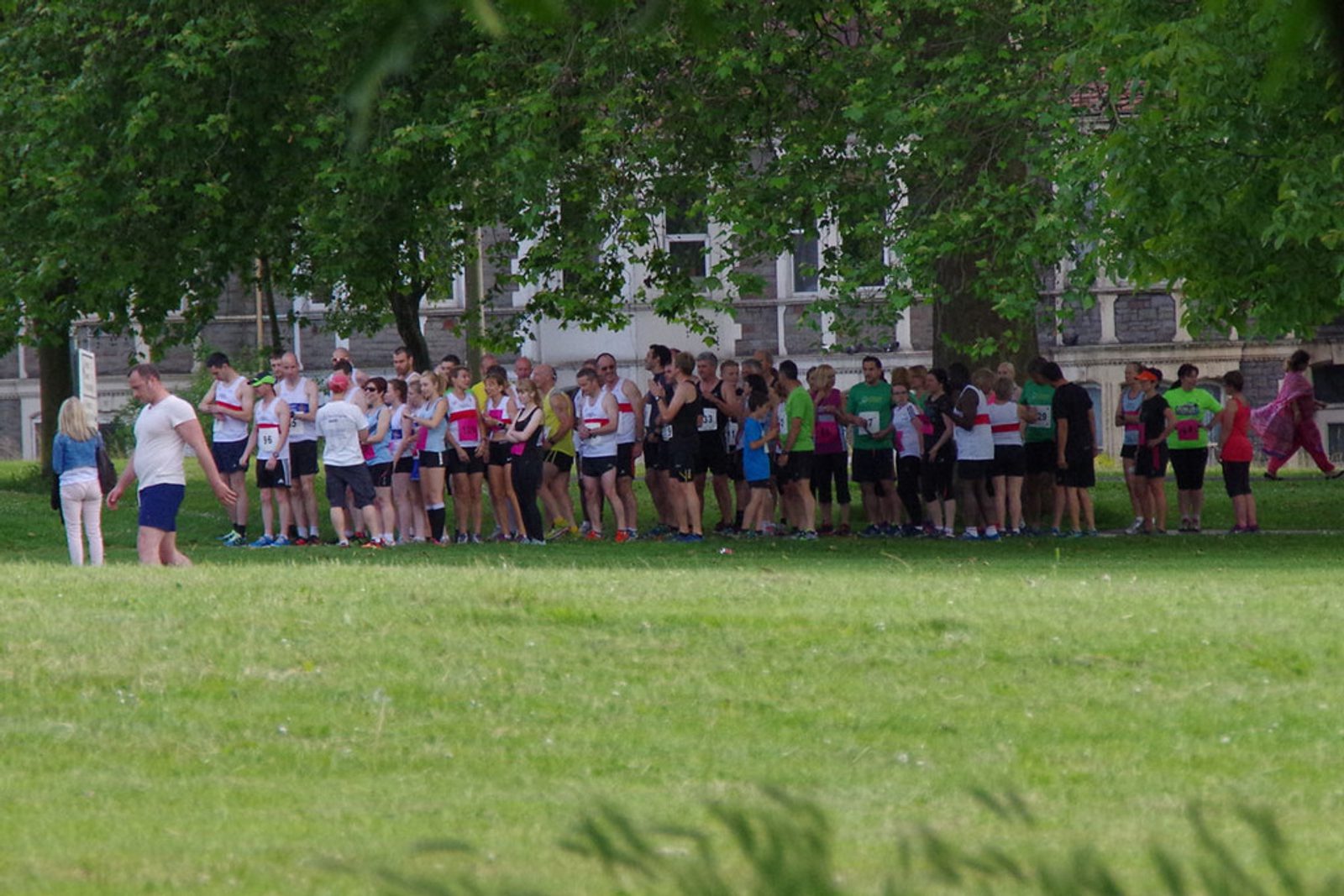 A group of runners, wearing a variety of colorful athletic clothing and numbered bibs, gather at the starting line in a park, preparing for a race. Trees and grass surround the area, and a few spectators are visible in the background.