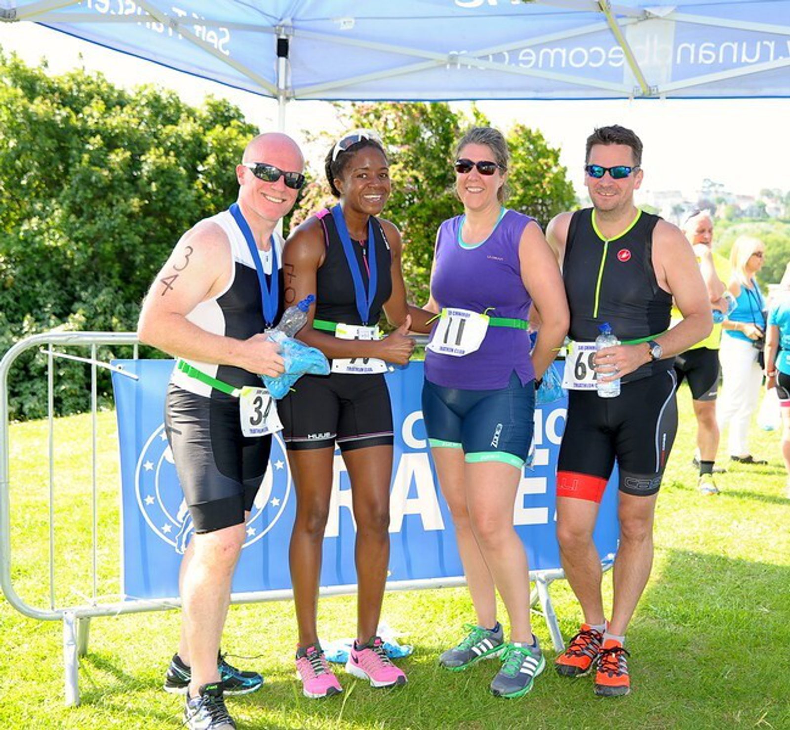 Four athletes, three wearing race numbers, pause for a photo after finishing a race, smiling and holding water bottles. They stand under a blue tent on a grassy area, with medals around their necks, in bright daylight. Green trees are visible in the background.