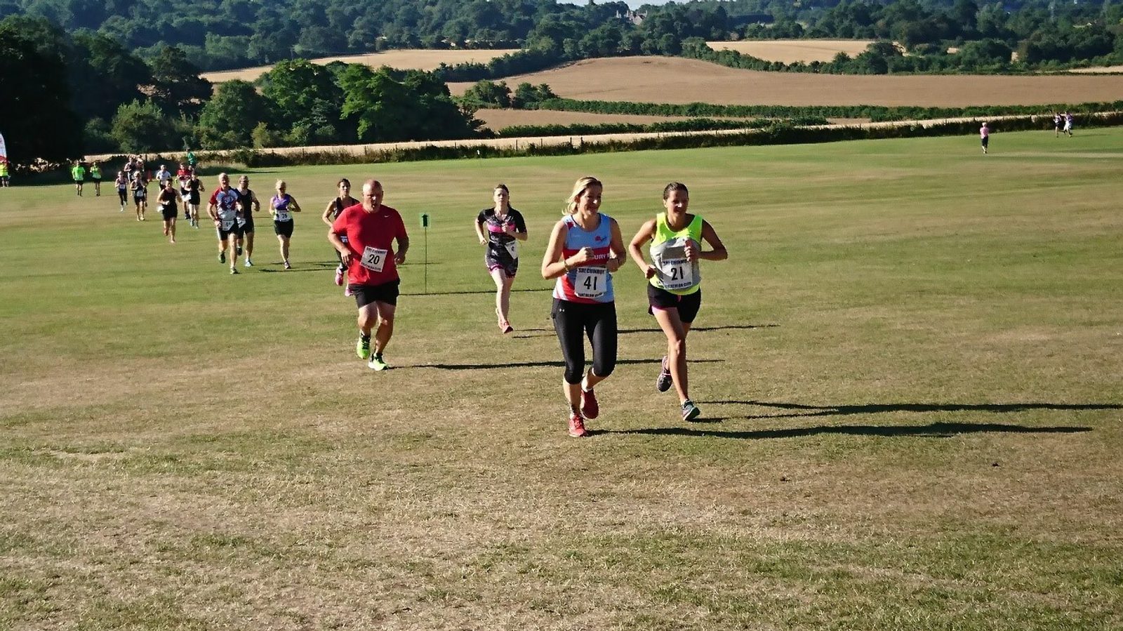 A group of runners participating in an outdoor race on a grassy field with rolling hills and trees in the background. The runners, both male and female, are wearing numbered bibs and athletic clothing as they move towards the camera. The sun is shining brightly.