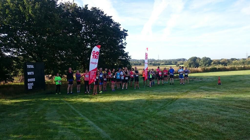 A group of runners line up at the starting point of a race on a grassy field. Visible in the background are trees, a clear sky, and a sign on the left displaying race information. Two red "GO TRI" banners are placed along the starting area.