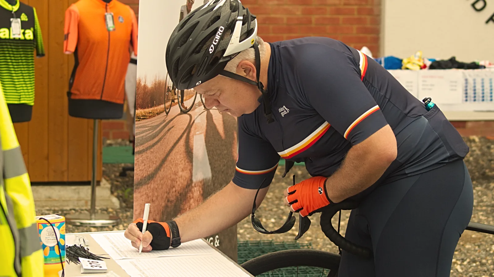 A cyclist, wearing a helmet and cycling gear, leans over to sign a sheet on a table. He is holding a pen in his right hand, and his left hand grips his bike’s handlebar. A brick wall and cycling gear are visible in the background.