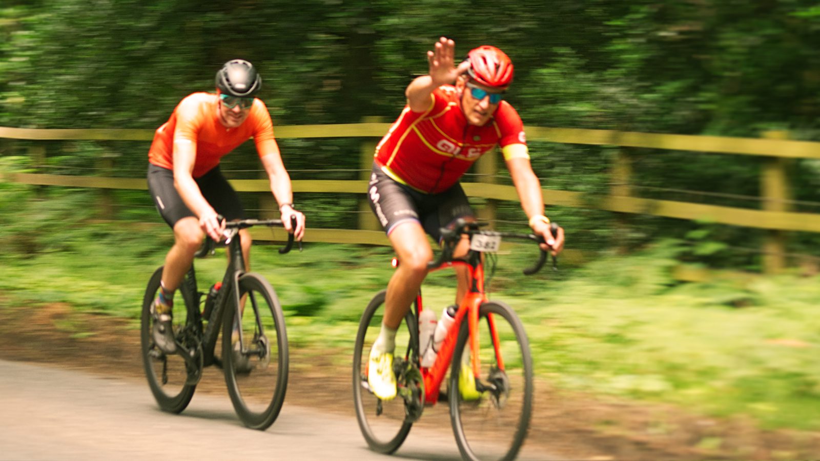 Two cyclists ride on a road beside a wooden fence. The cyclist in front is wearing a red jersey and is raising a hand, while the cyclist behind is in an orange jersey, both wearing helmets. The background is a blur of green vegetation, indicating movement.