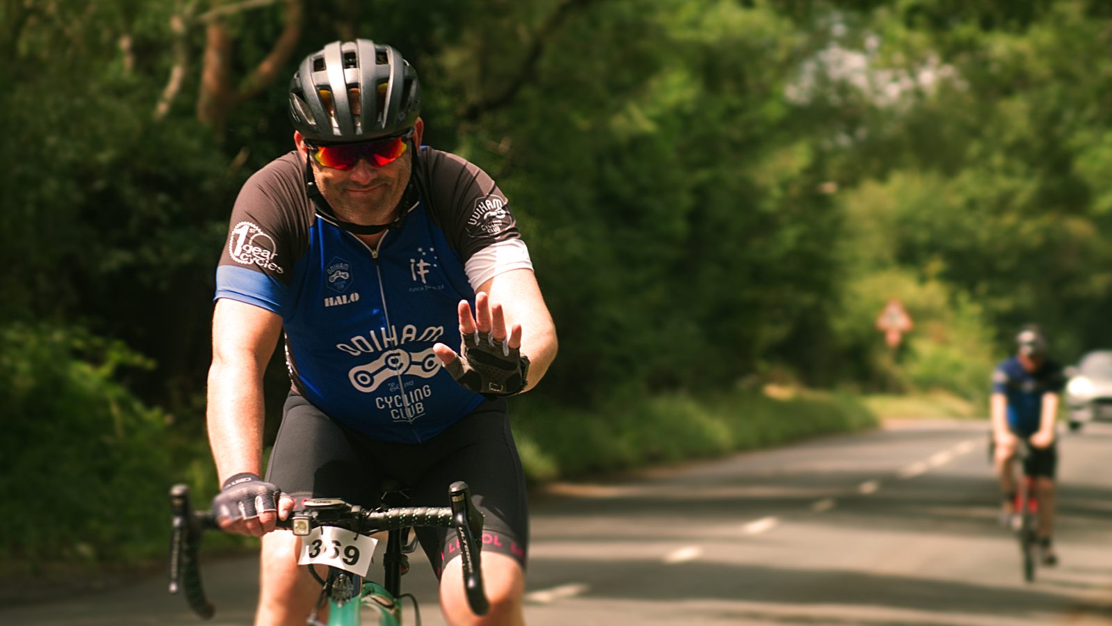 A cyclist wearing a black helmet and a blue and black cycling jersey is riding a bike on a road lined with greenery. He is smiling and waving at the camera with his left hand. Another cyclist is seen in the background. The cyclist has the number 139 on his bike.