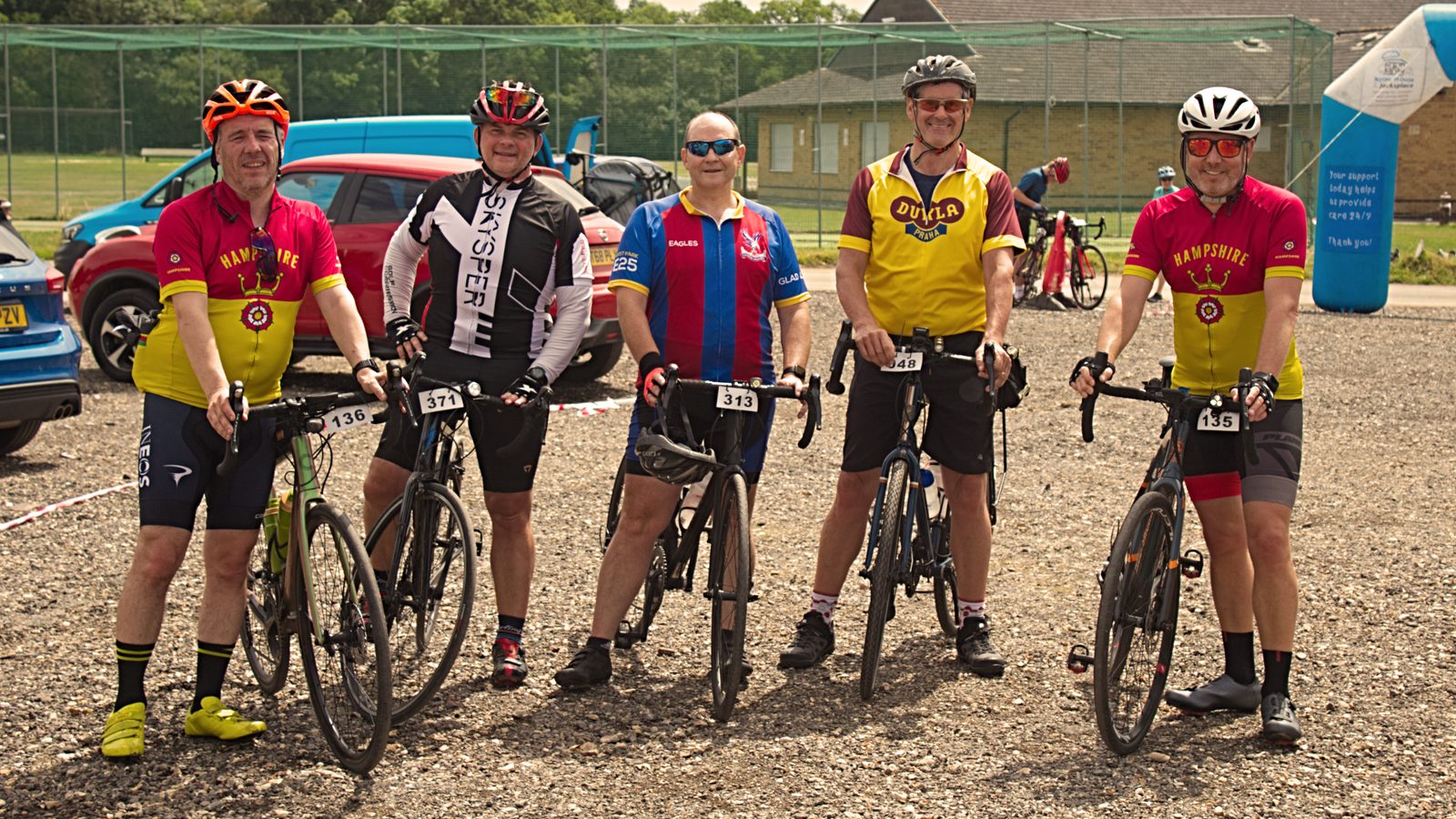 A group of five cyclists dressed in colorful cycling attire pose with their bicycles in an outdoor setting. They stand on a gravel surface, with cars and buildings visible in the background. All five individuals are wearing helmets and sunglasses, preparing for a ride.