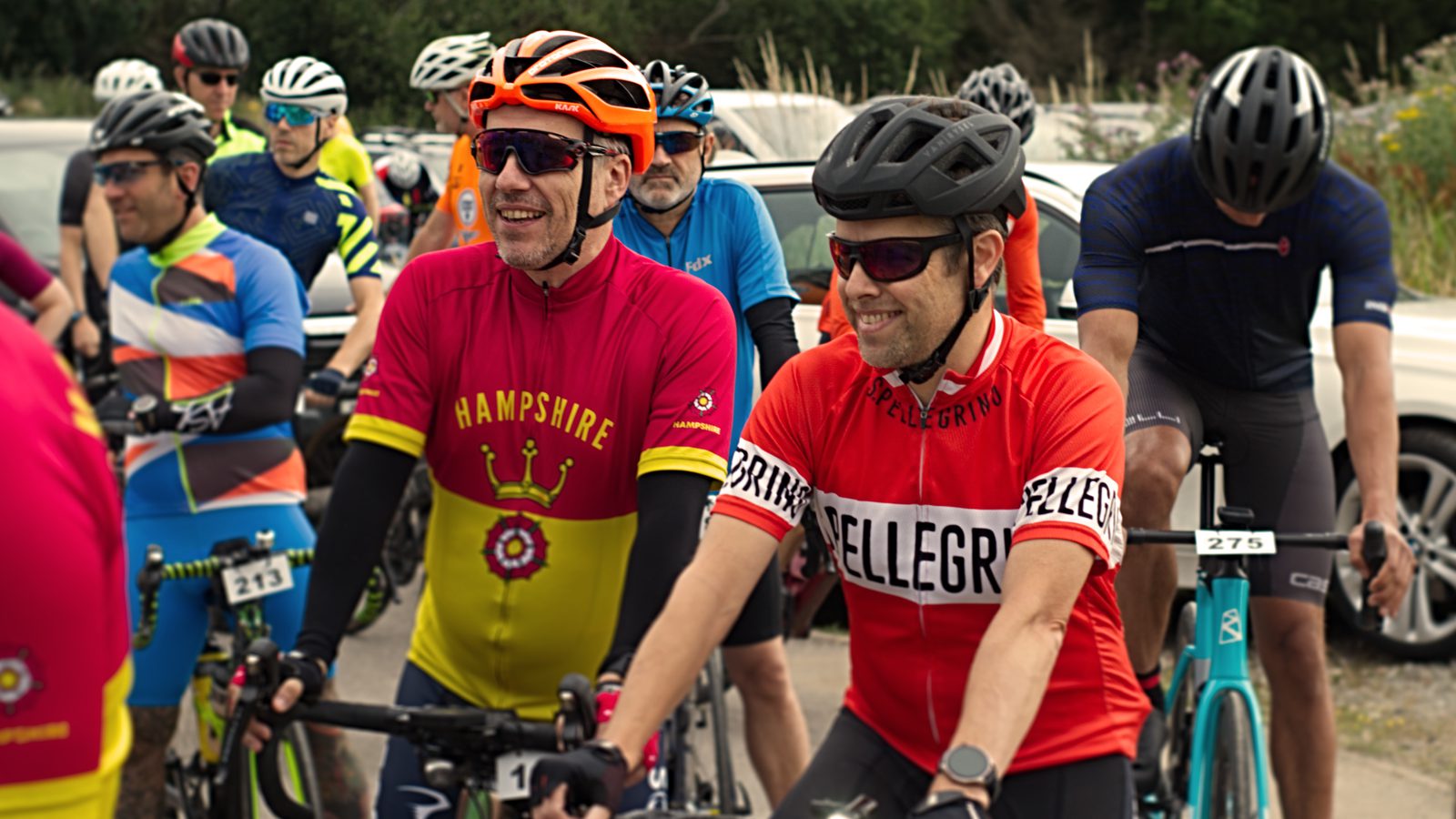 A group of cyclists dressed in colorful jerseys and helmets gather at the starting line of a race. Two men in the foreground are smiling and chatting, one in a yellow and red "Hampshire" jersey, the other in a red "Bellegrove" jersey. Bicycles and cars are visible in the background.