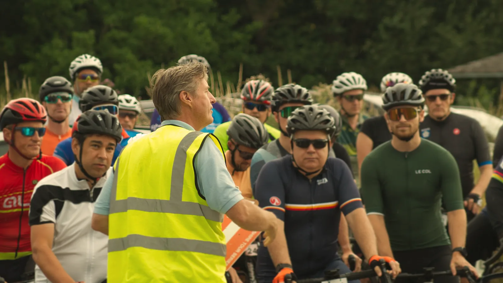 A man in a yellow reflective vest addresses a group of cyclists gathered around him. The cyclists, wearing various colorful jerseys and helmets, listen attentively. The background shows greenery, suggesting an outdoor setting.