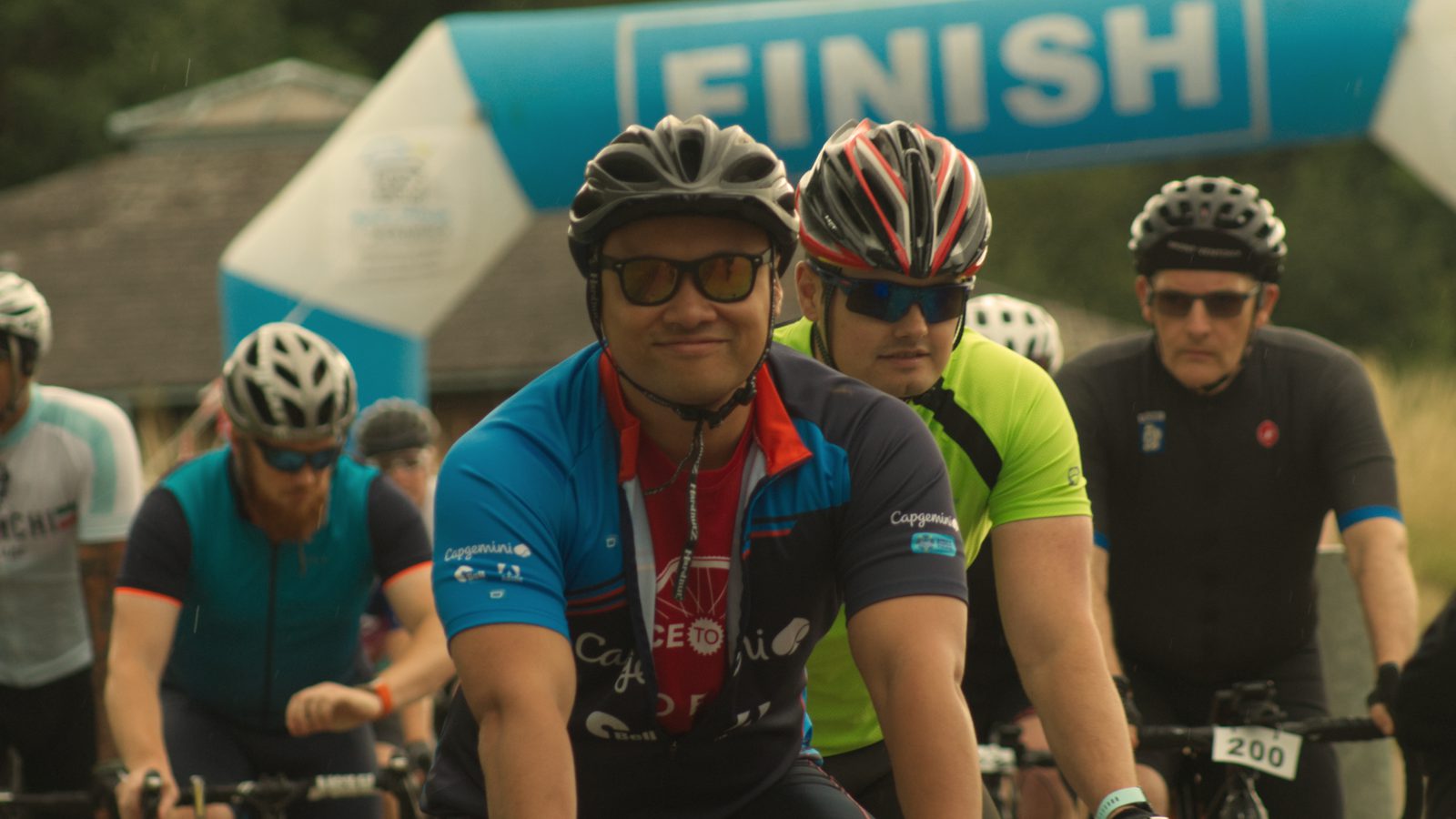Cyclists cross the finish line during a race. The person in the foreground wears a blue and red jersey, sunglasses, and a helmet, smiling at the camera. Behind him, other cyclists in bright jerseys and helmets follow. A large "FINISH" banner is visible above them.