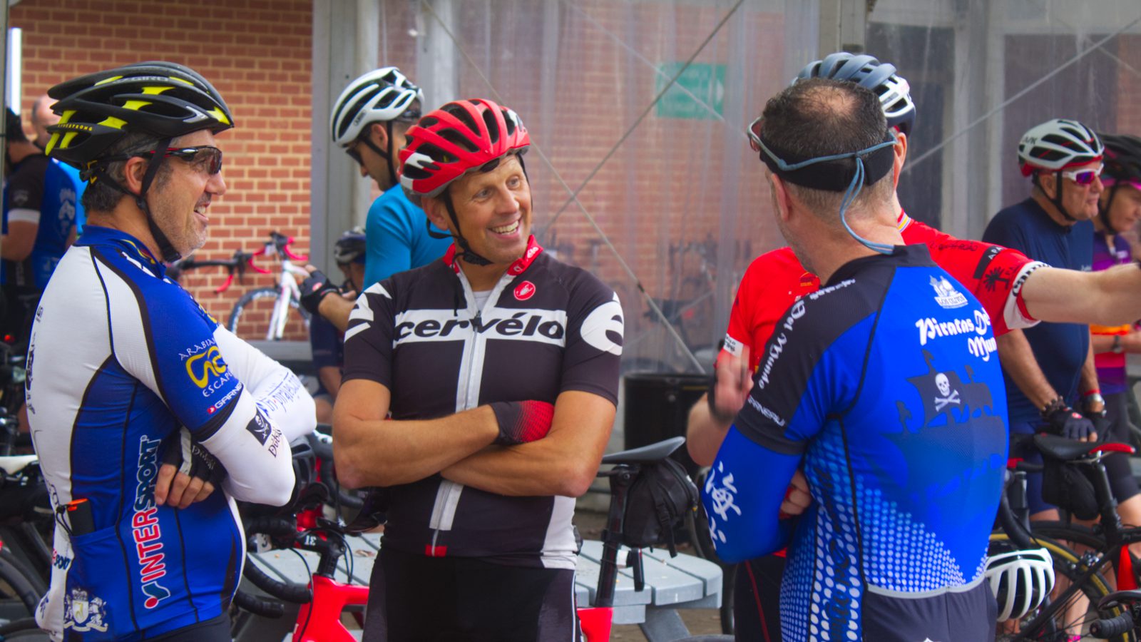 A group of cyclists in colorful jerseys and helmets stand together, conversing and smiling. Bicycles are parked in the background. The scene appears to be at a gathering point before or after a cycling event. Brick and glass walls are also visible.