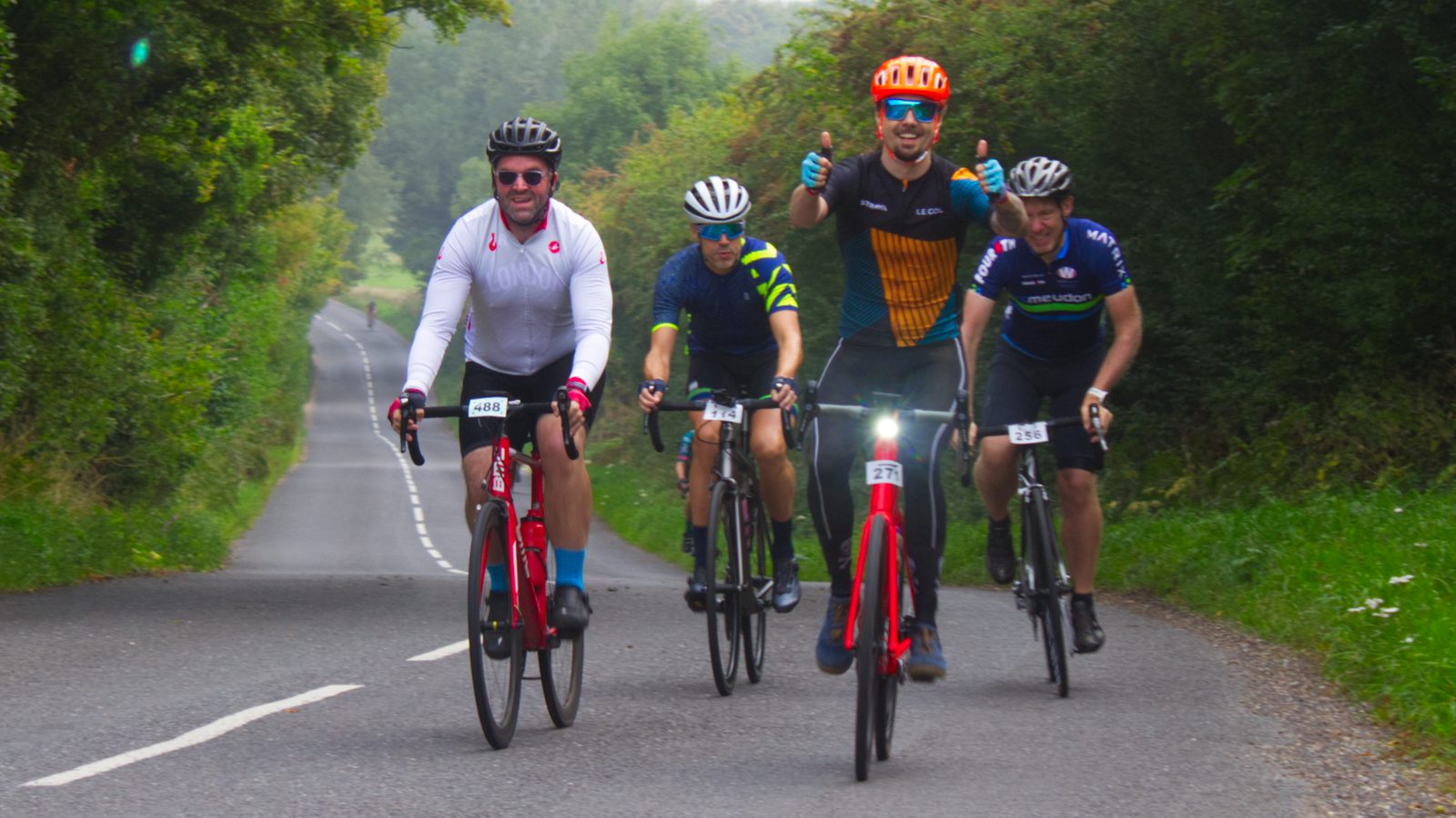 Four cyclists are riding on a two-lane road lined with green foliage. The central cyclist gives a thumbs-up while wearing an orange helmet and black and yellow jersey. The others wear various cycling attire, and the road stretches into the distance behind them.